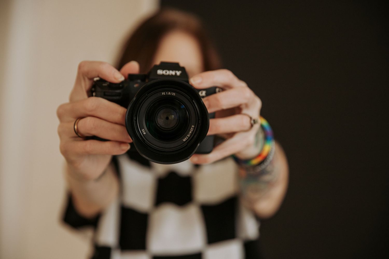 A person in a checkered shirt holds a Sony DSLR camera up to their face, wearing colorful bracelets.