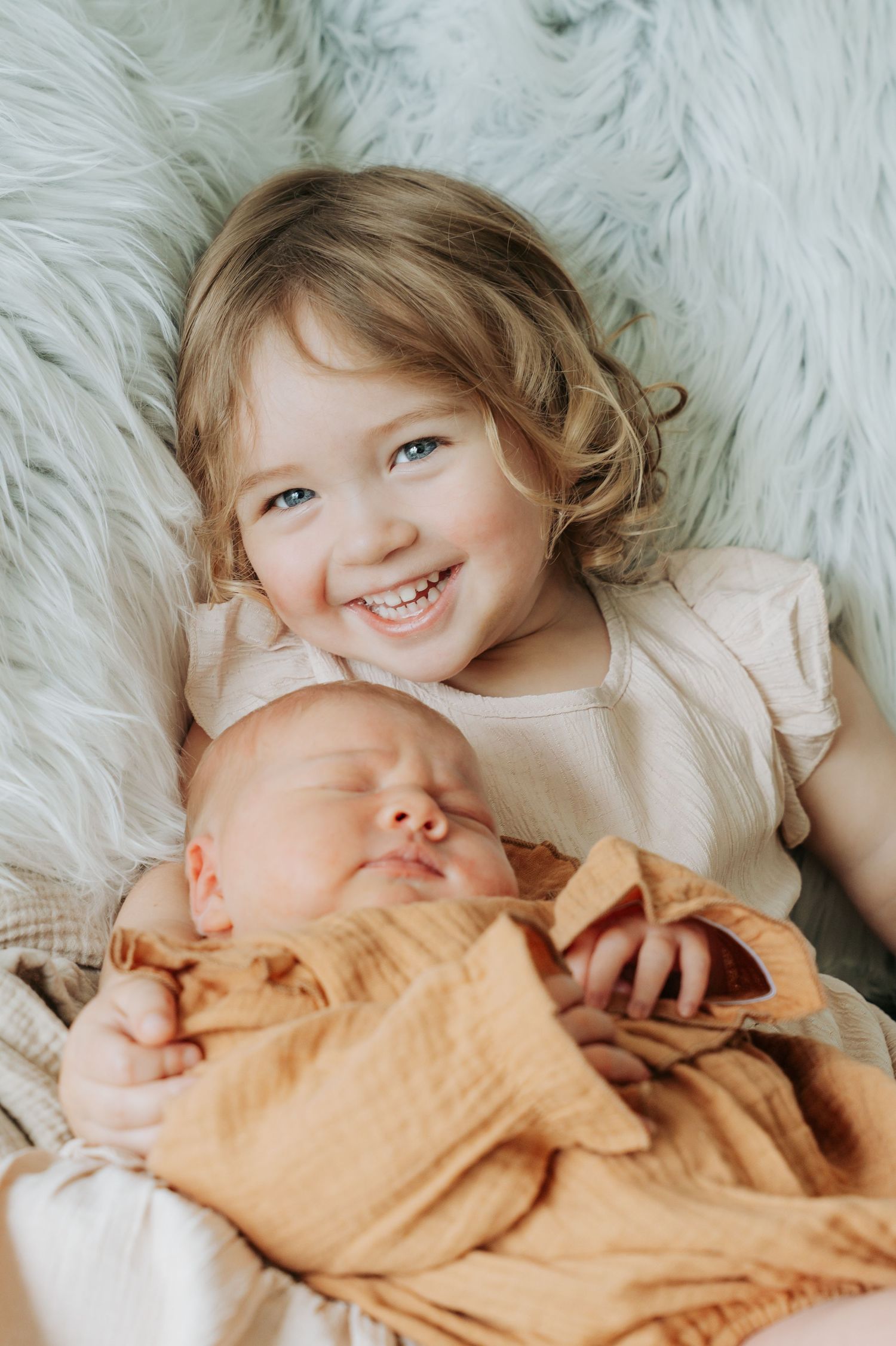 Smiling toddler girl with curly hair lying next to a sleeping newborn baby wrapped in orange muslin.
