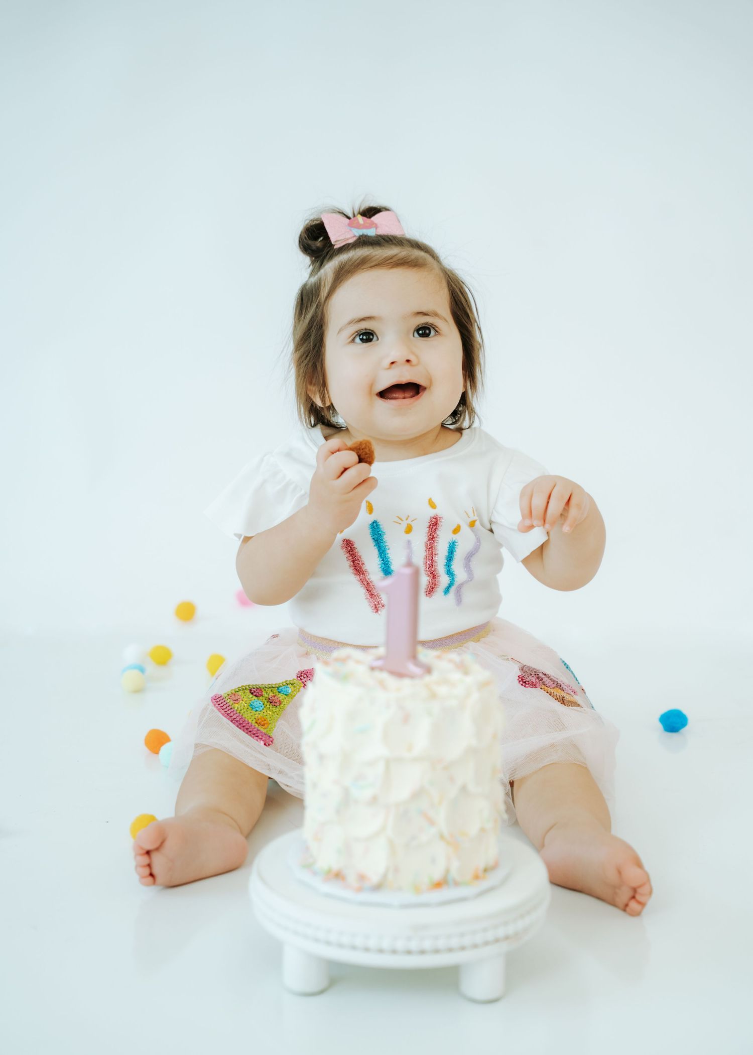 Adorable baby girl in white dress celebrates first birthday with colorful cake smash setup.