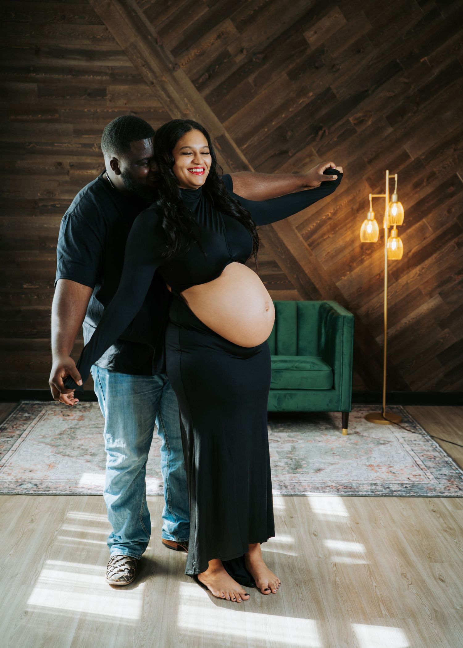 Pregnant couple poses in rustic wood-paneled room, woman in black maternity dress cradling baby bump.