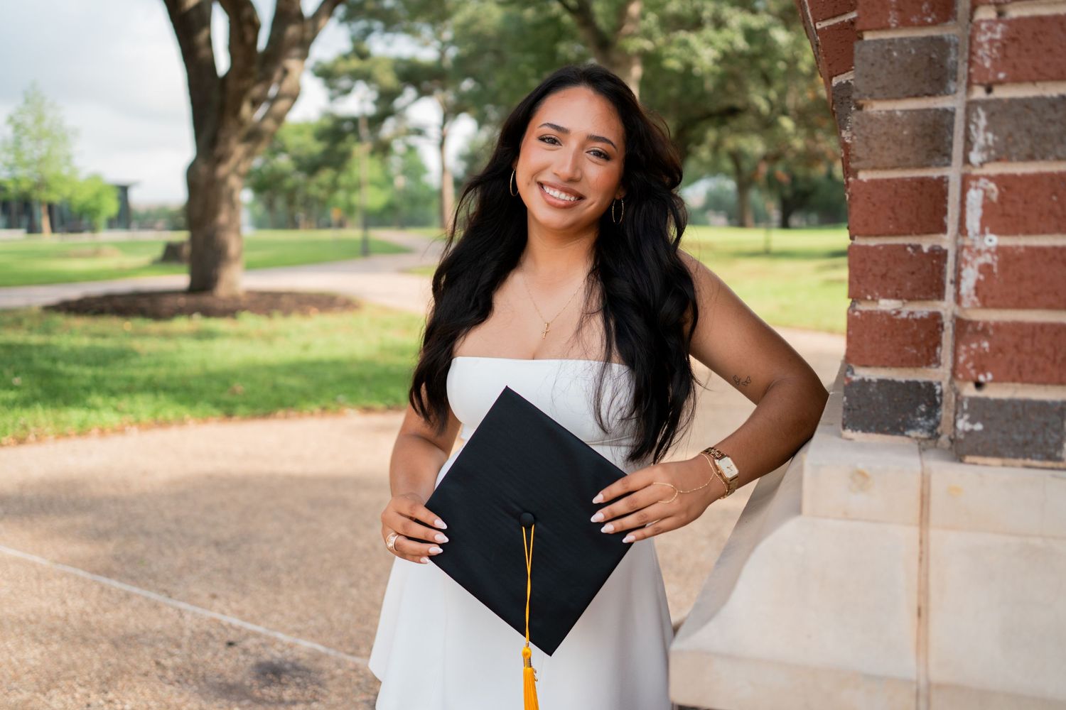 Smiling young woman in white dress holding graduation cap with gold tassel outdoors on campus.