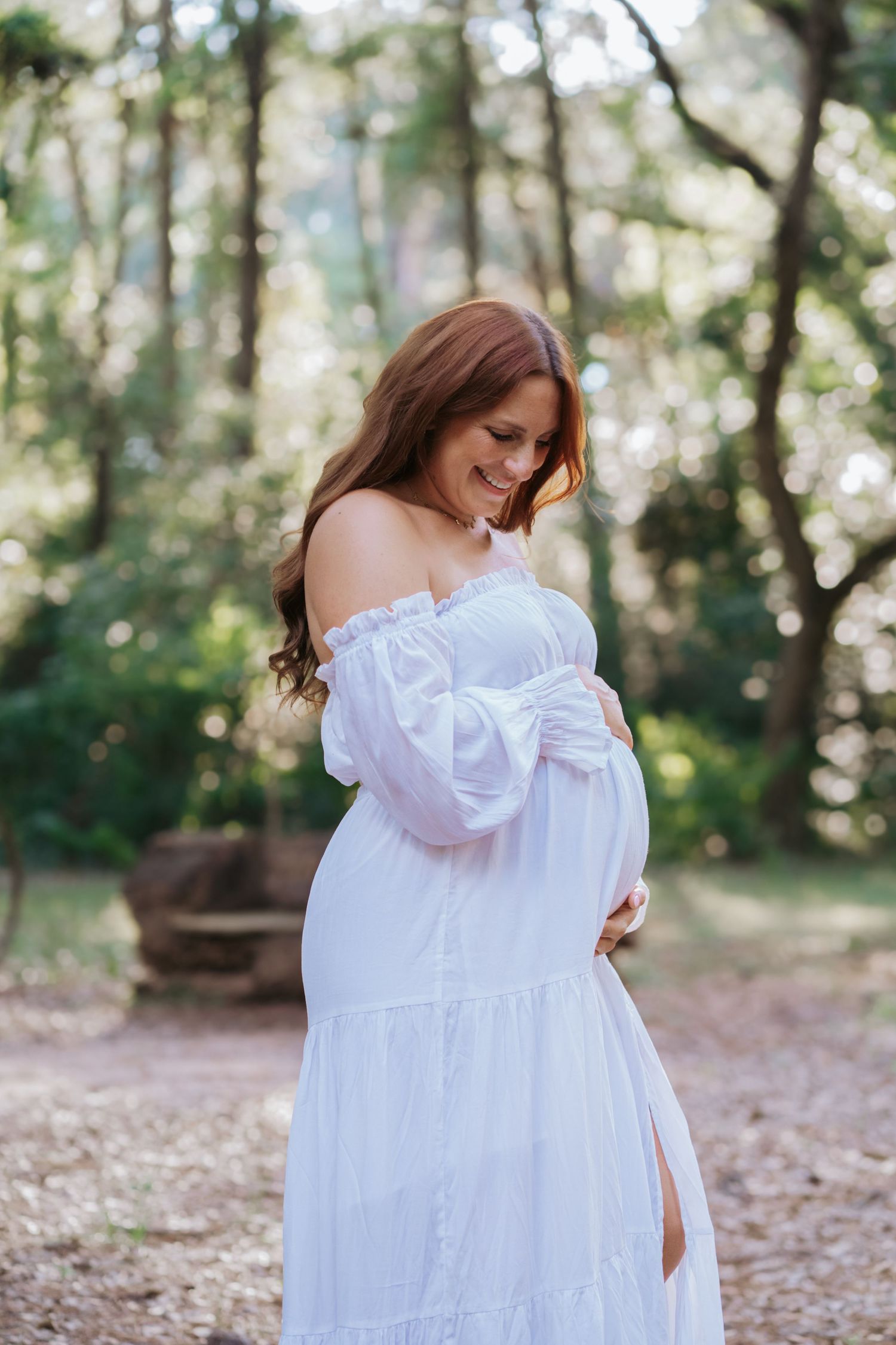 Glowing pregnant woman in white off-shoulder dress cradles baby bump in sunlit forest setting.