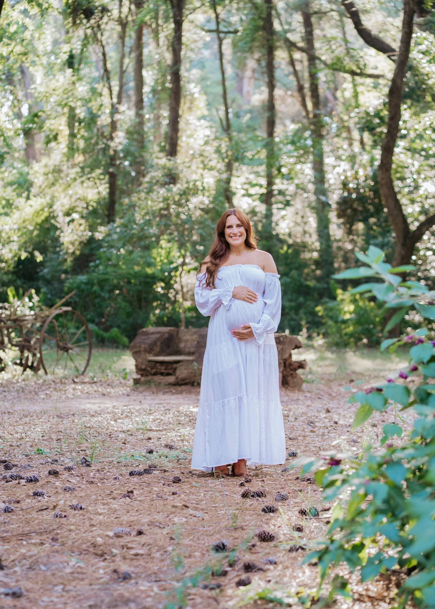 Smiling pregnant woman in white off-shoulder dress poses on forest path during maternity session.