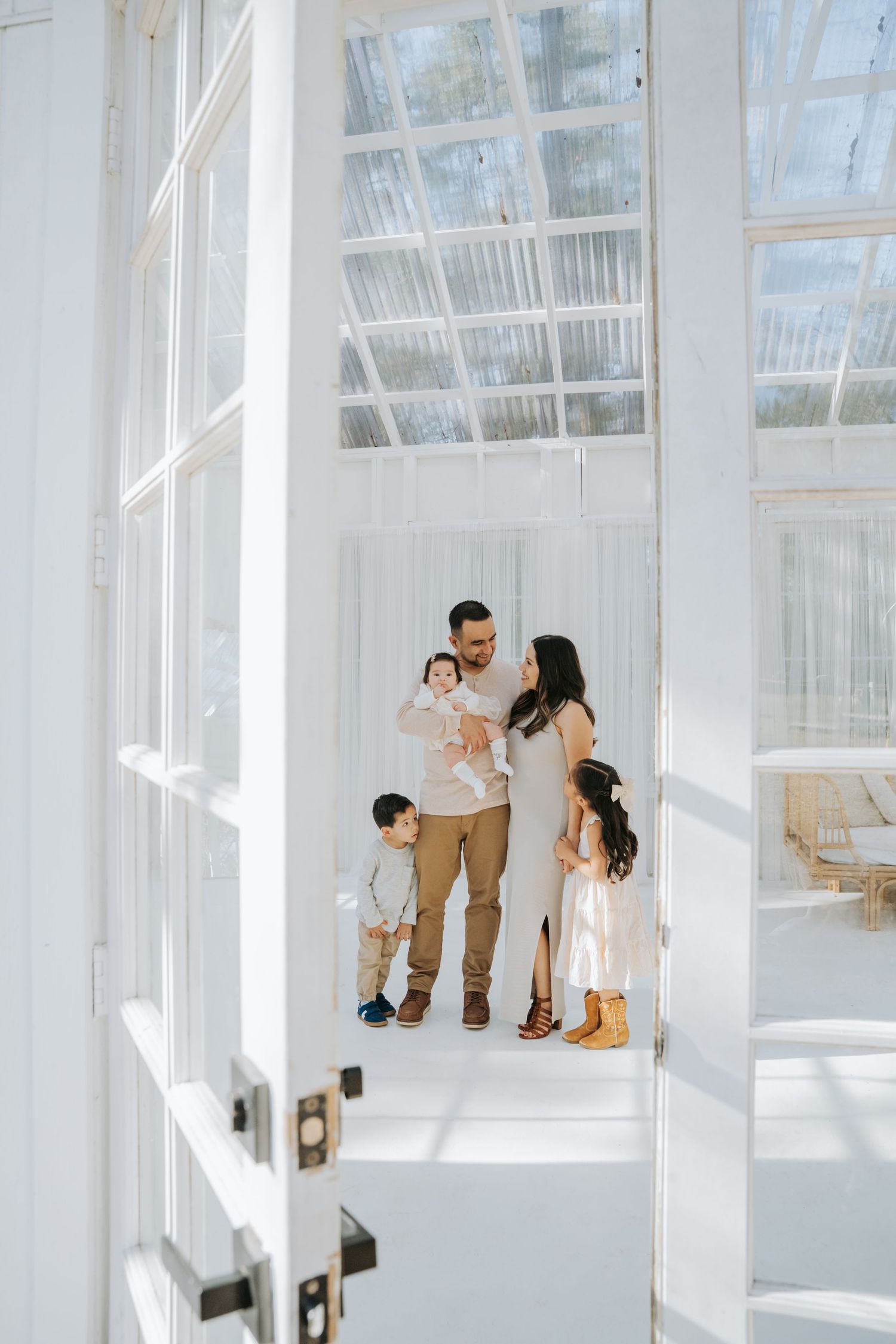Family of four poses in a bright white greenhouse, parents holding baby while toddler stands beside them.
