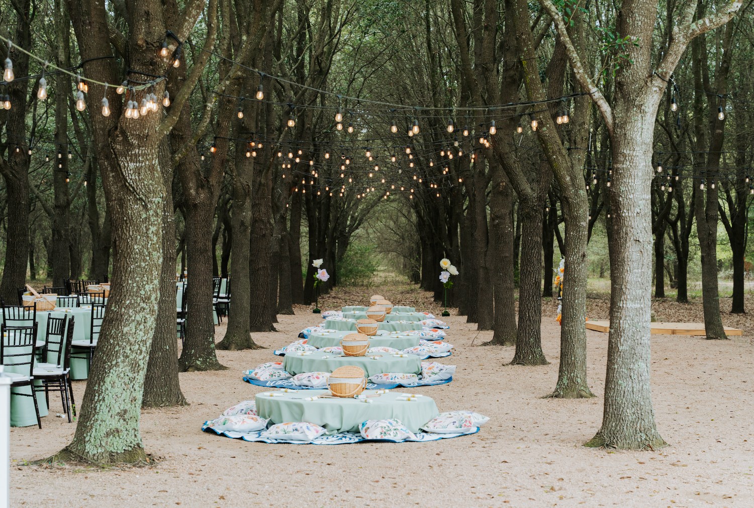 Enchanting outdoor wedding reception with string lights, round tables, and lush tree-lined pathway in a forest setting.