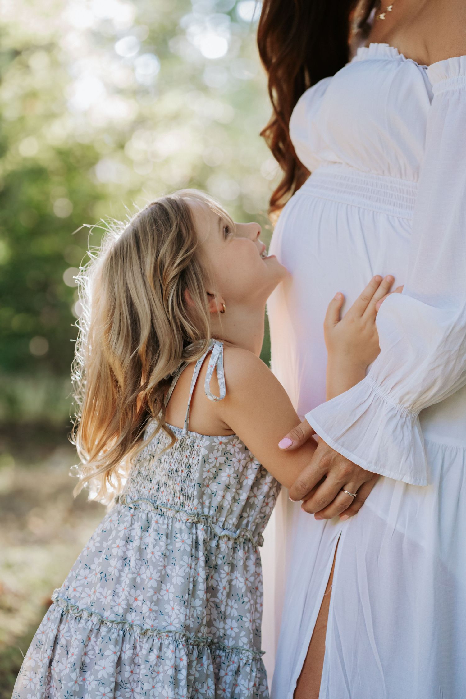 Young blonde girl in floral dress smiling up at her pregnant mothers baby bump outdoors.