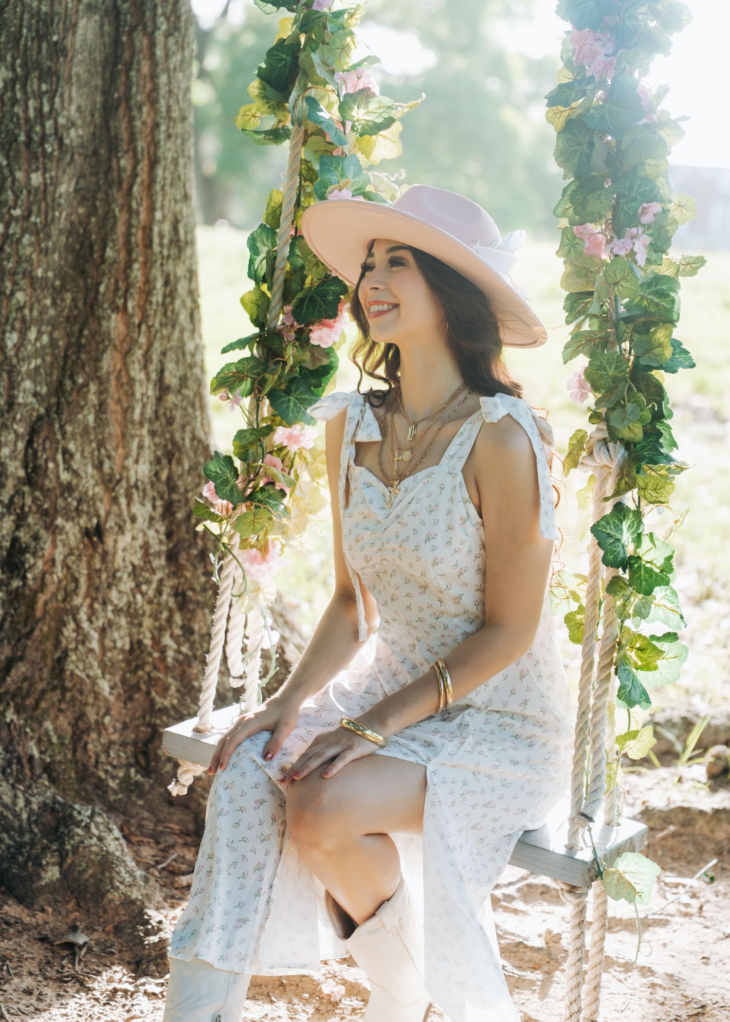 Woman in white floral dress and pink hat sitting on a flower-adorned rope swing by a tree.