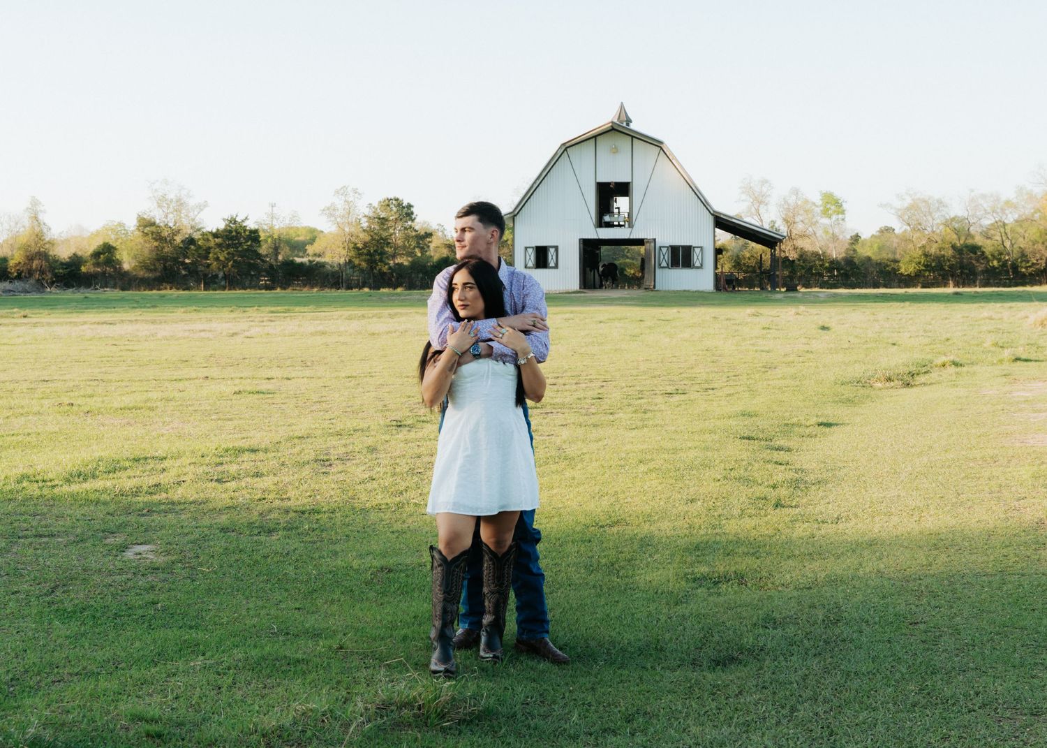 Couple embracing in front of a white barn on a green field during golden hour at a farm engagement shoot.