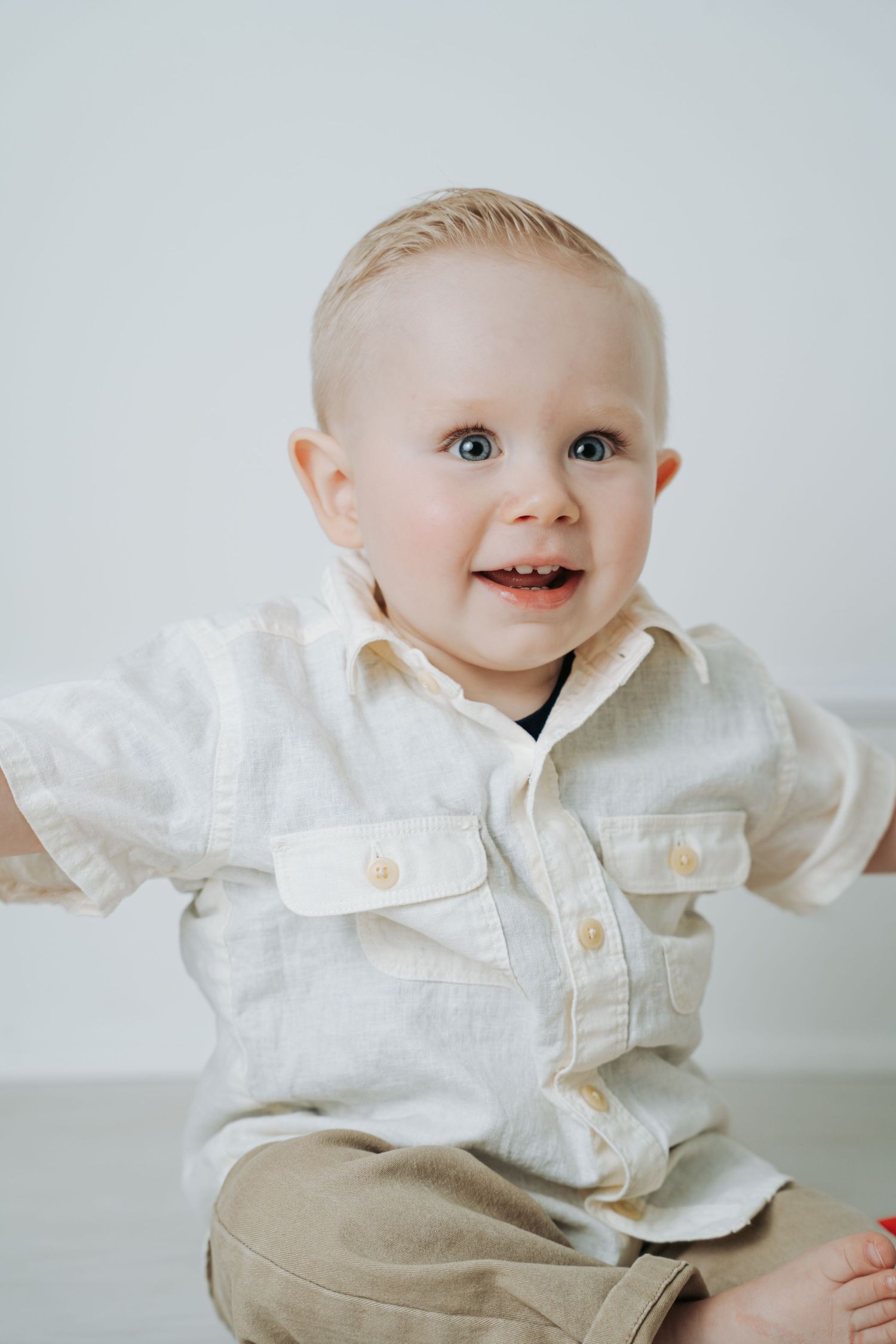 Smiling blond toddler boy with blue eyes wearing a white linen shirt and tan pants sitting against a white background.