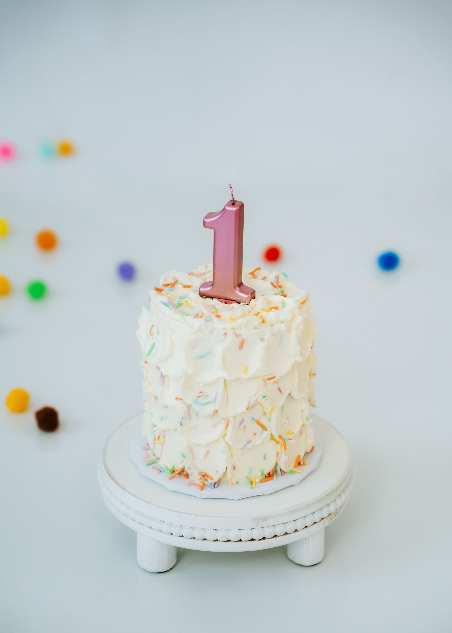 A white first birthday smash cake with pink number one candle surrounded by colorful pom poms.