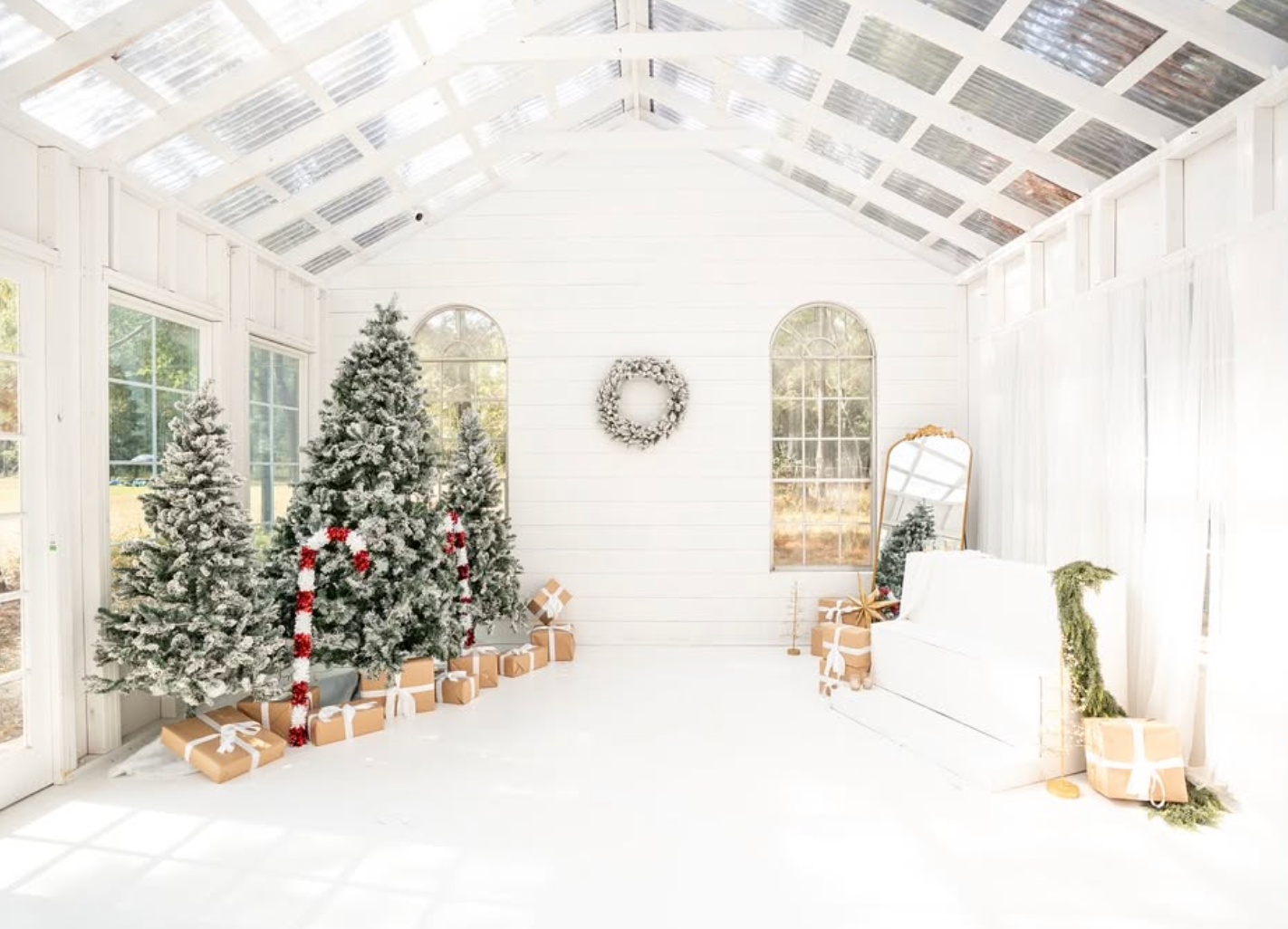 Bright white greenhouse decorated for Christmas with snow-flocked trees, wrapped gifts, and a festive wreath on the wall.