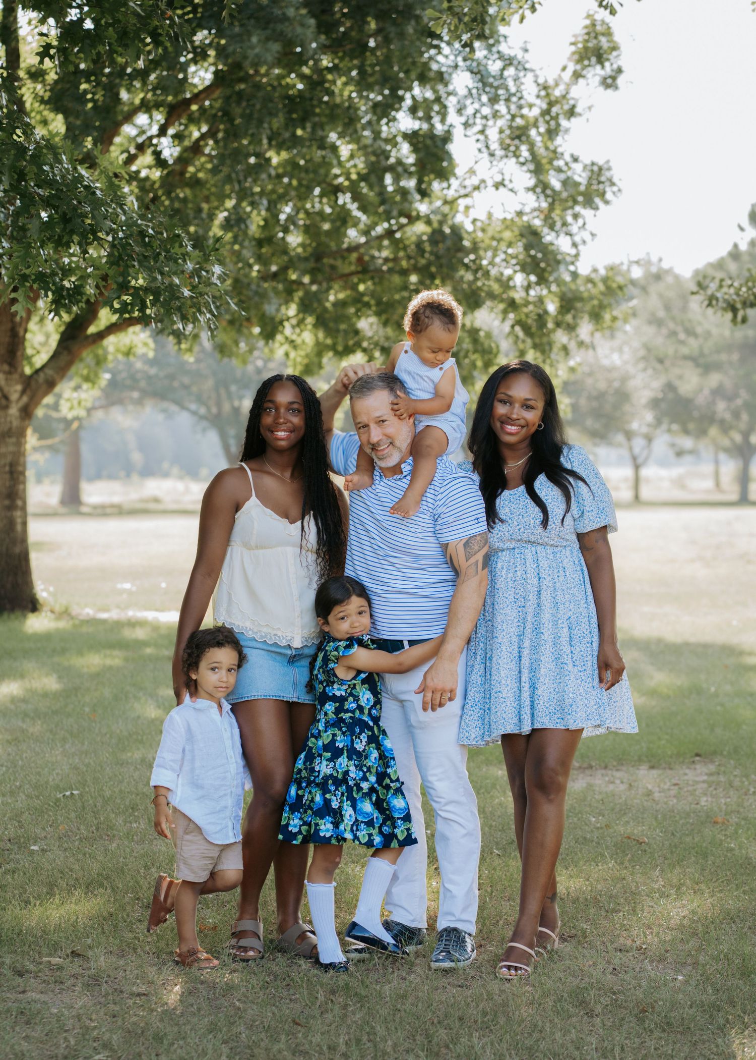 Blended family of six posing together under large tree in sunny park during outdoor session.