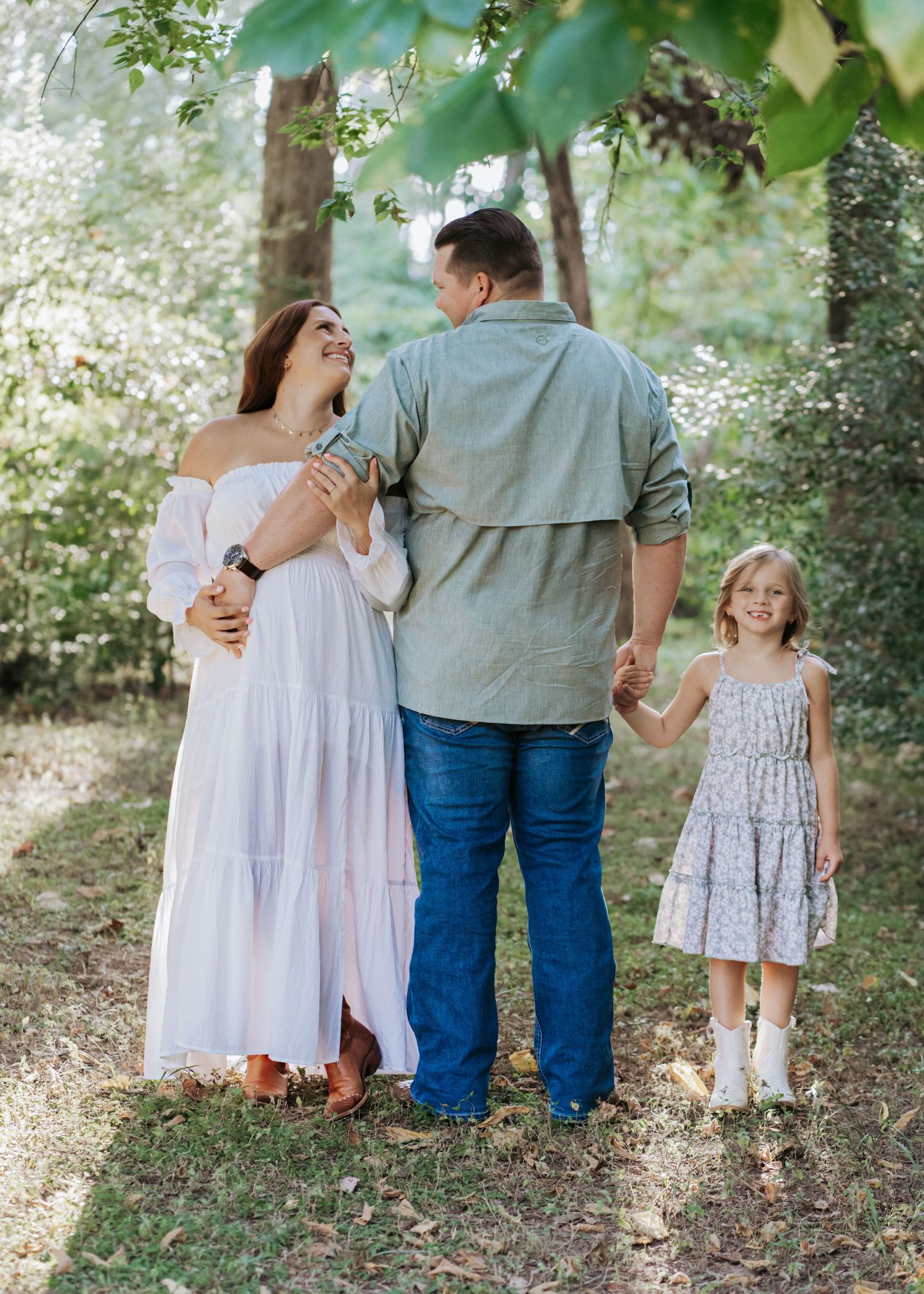 Family of three walks through lush green forest, pregnant mother in white dress holding hands with daughter.