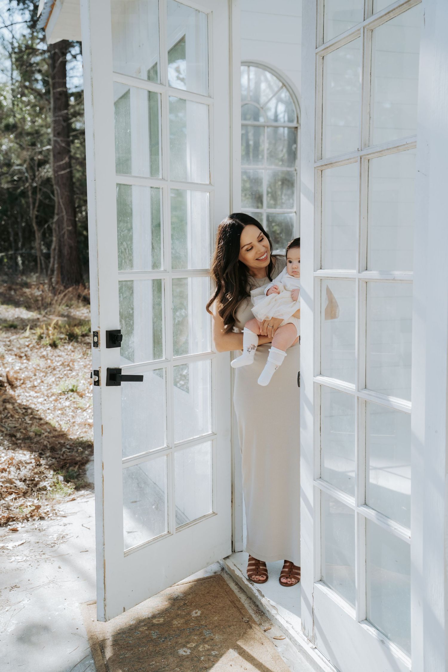A brunette mother in a white dress holds her baby while standing in elegant white French doors.