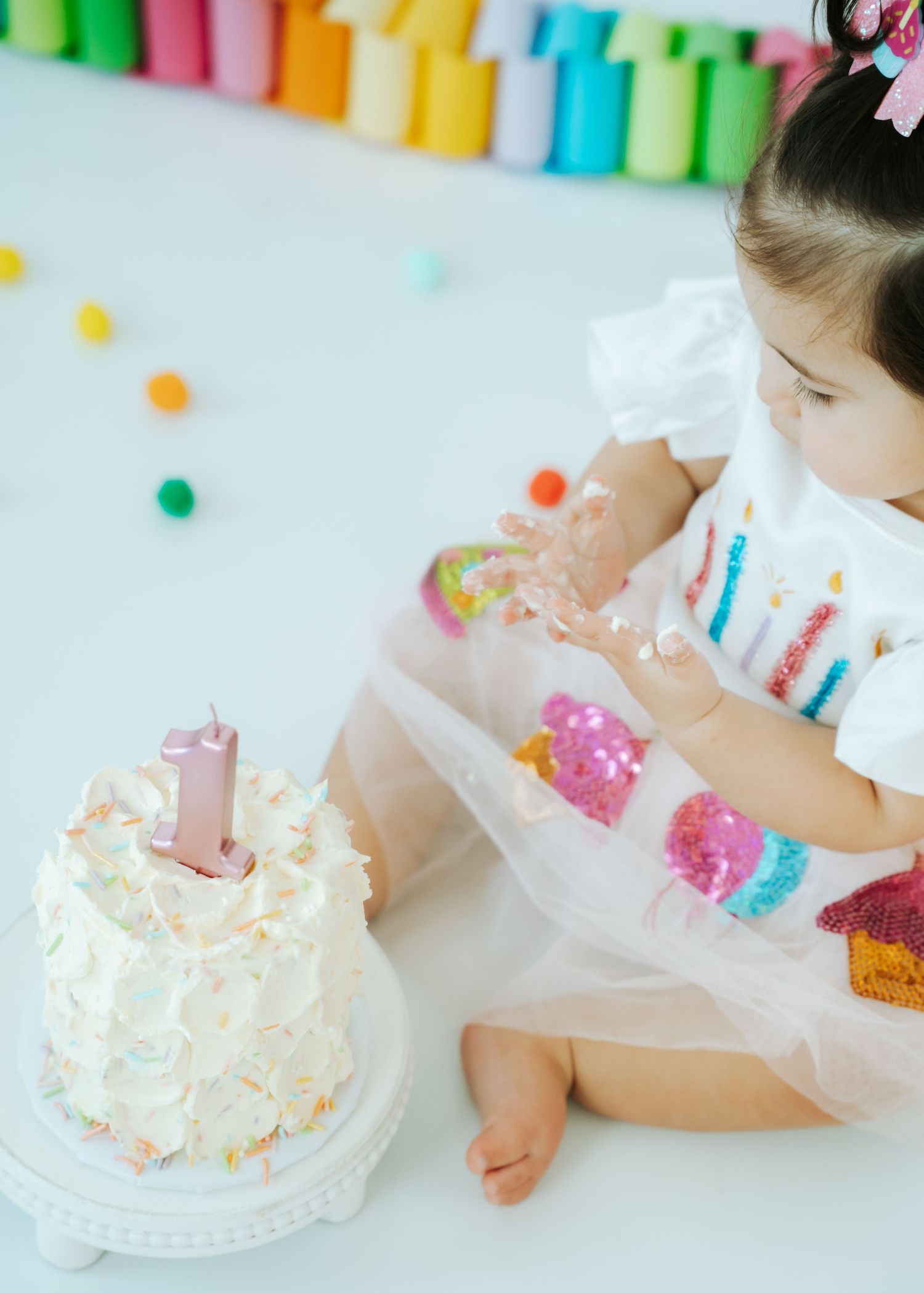 Baby girl in white tutu dress explores her first birthday smash cake with colorful sprinkles.