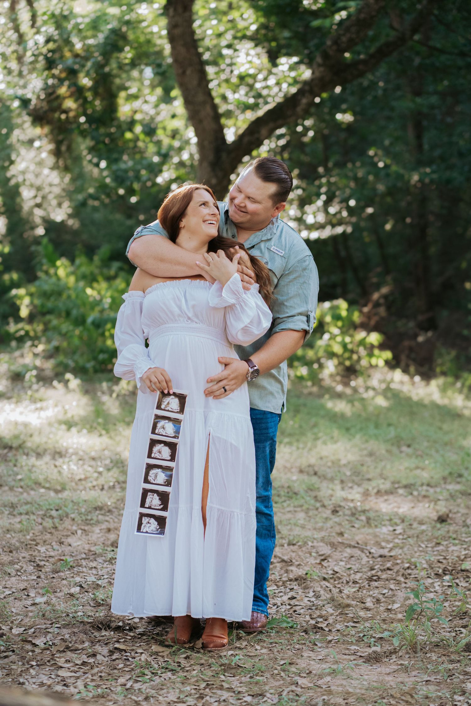 Smiling pregnant couple embraces outdoors holding ultrasound photos, woman in white off-shoulder dress.