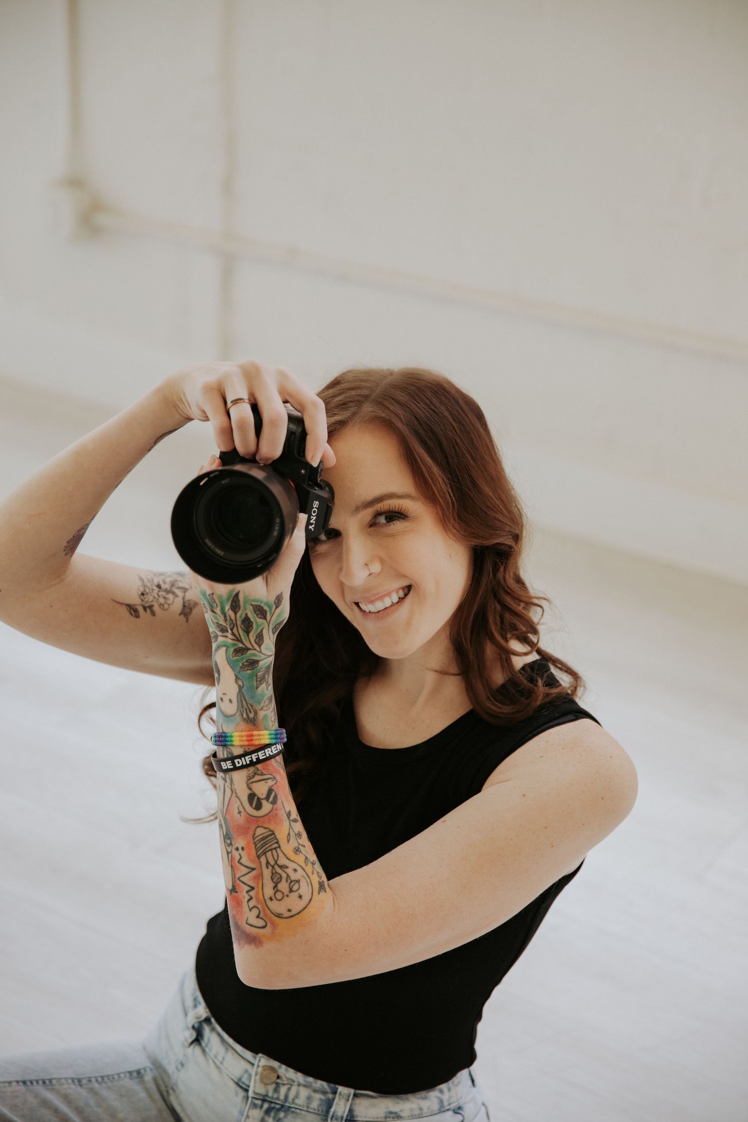 Smiling female photographer with tattoos holds DSLR camera in bright studio setting.