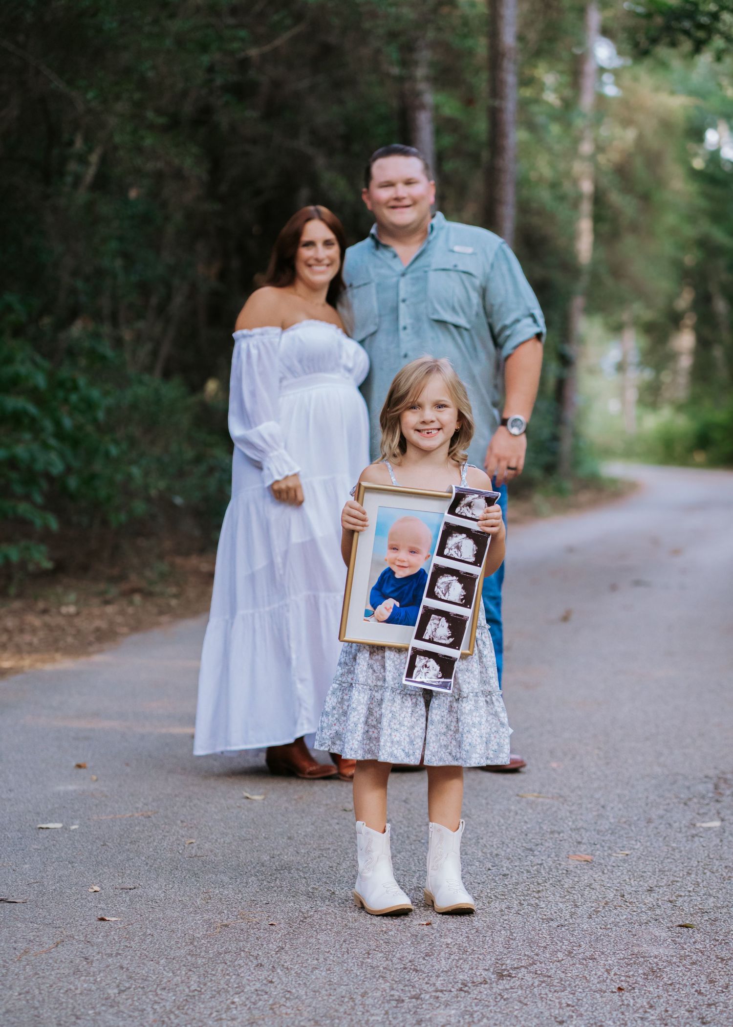 Pregnant couple standing on a tree-lined path while their daughter holds a framed sonogram photo.