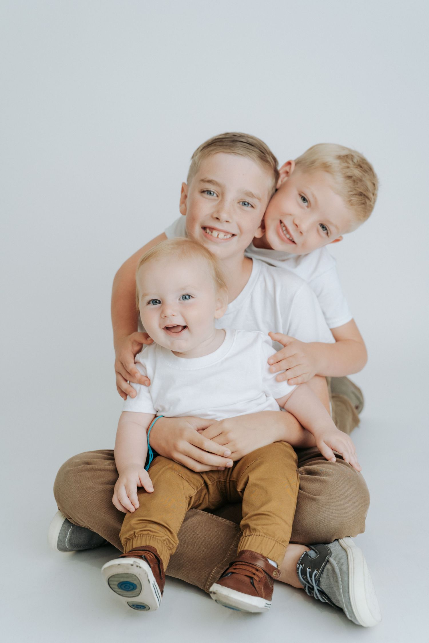 Three brothers pose together on white background, two older boys hugging baby sibling in matching white shirts.