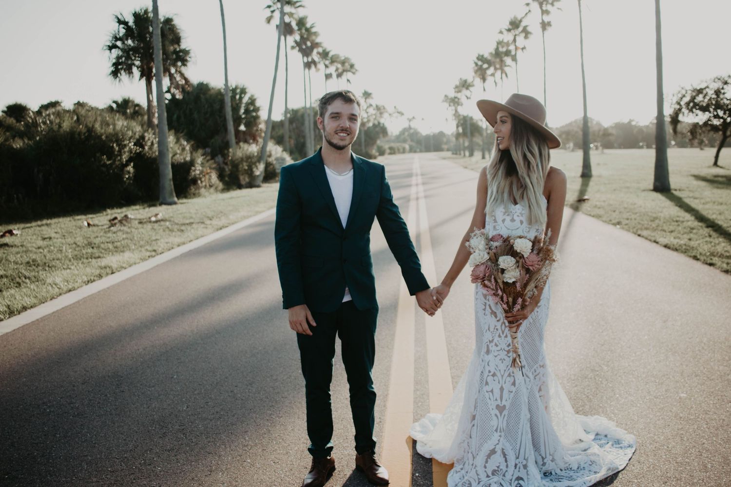 Bride and groom holding hands on a palm-lined road, bride wearing lace gown and boho hat, groom in navy suit.