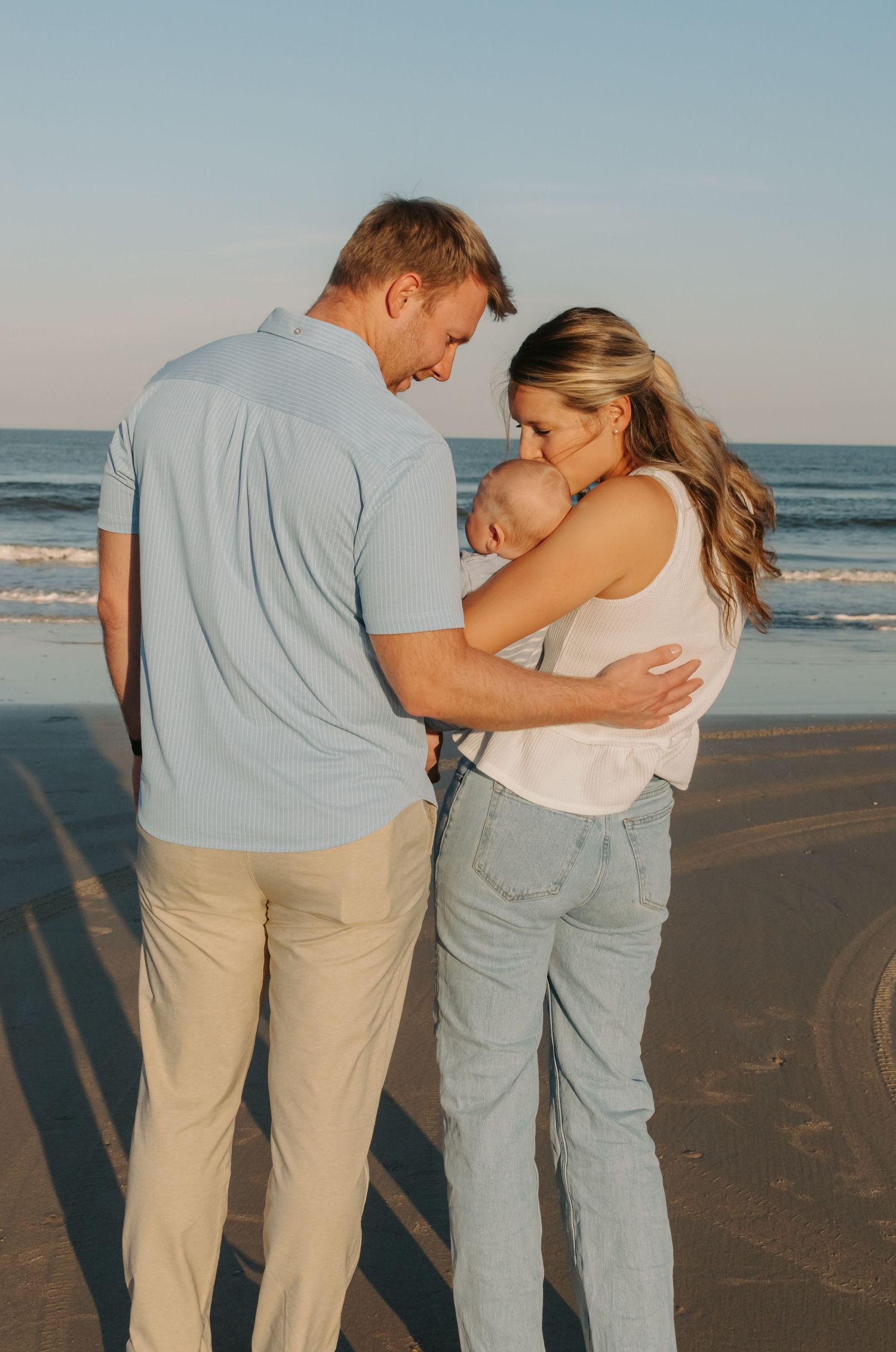 New parents tenderly holding newborn baby together on sandy beach at sunset.