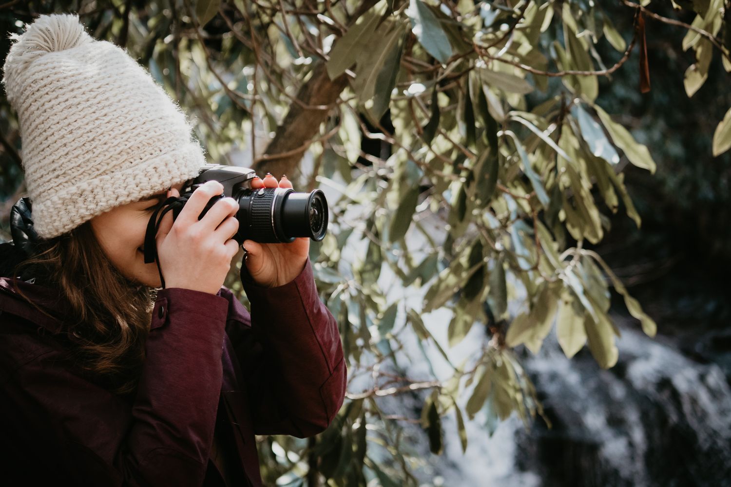Young woman in beige knit hat photographing nature with DSLR camera among green leafy branches outdoors.