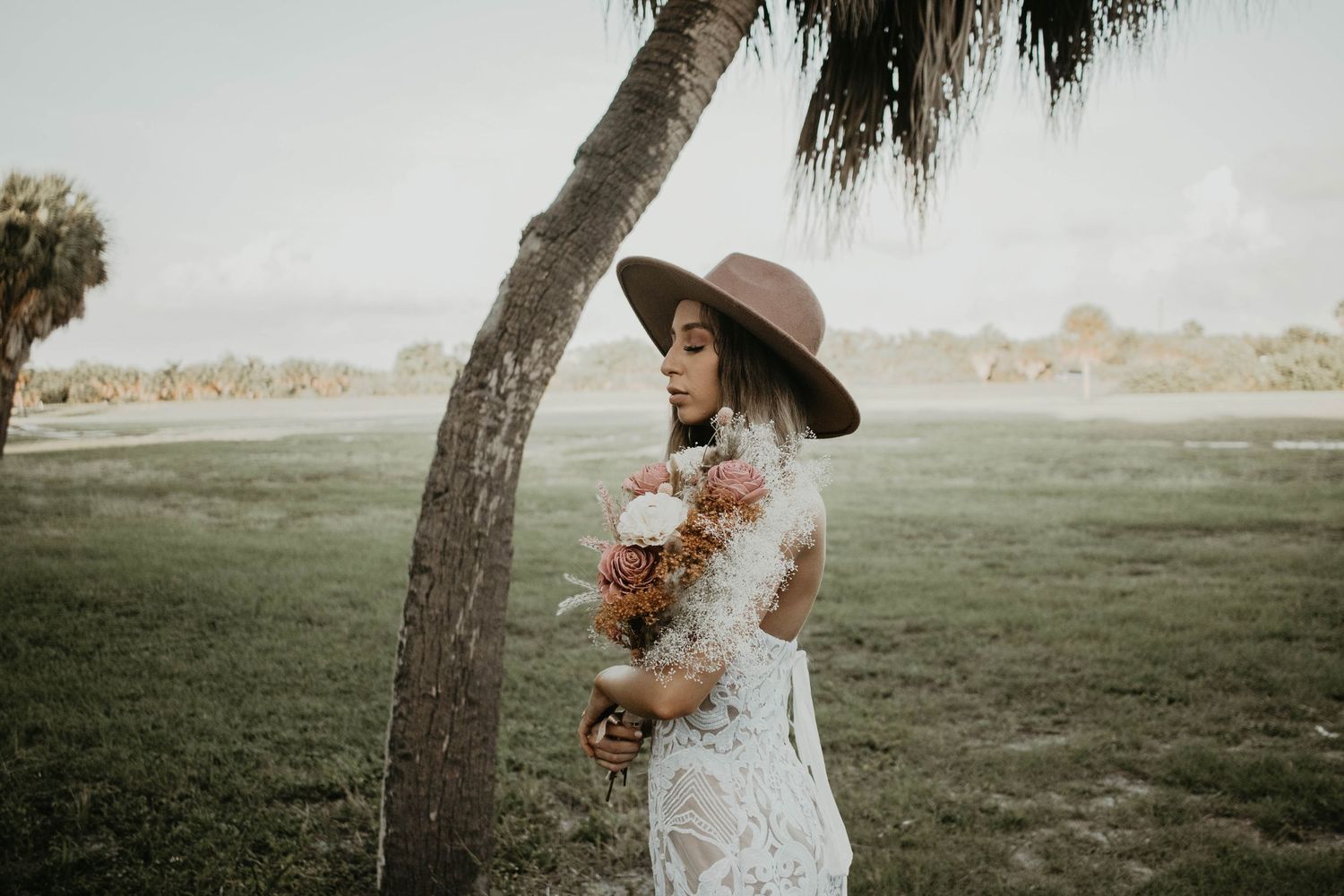 Bride in lace gown and brown wide-brim hat holds floral bouquet beside a palm tree in a lush green field.