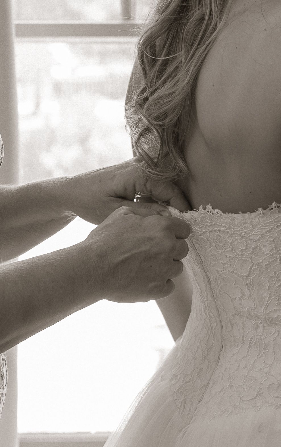 Black and white close-up of hands fastening lace wedding dress on bride near bright window.