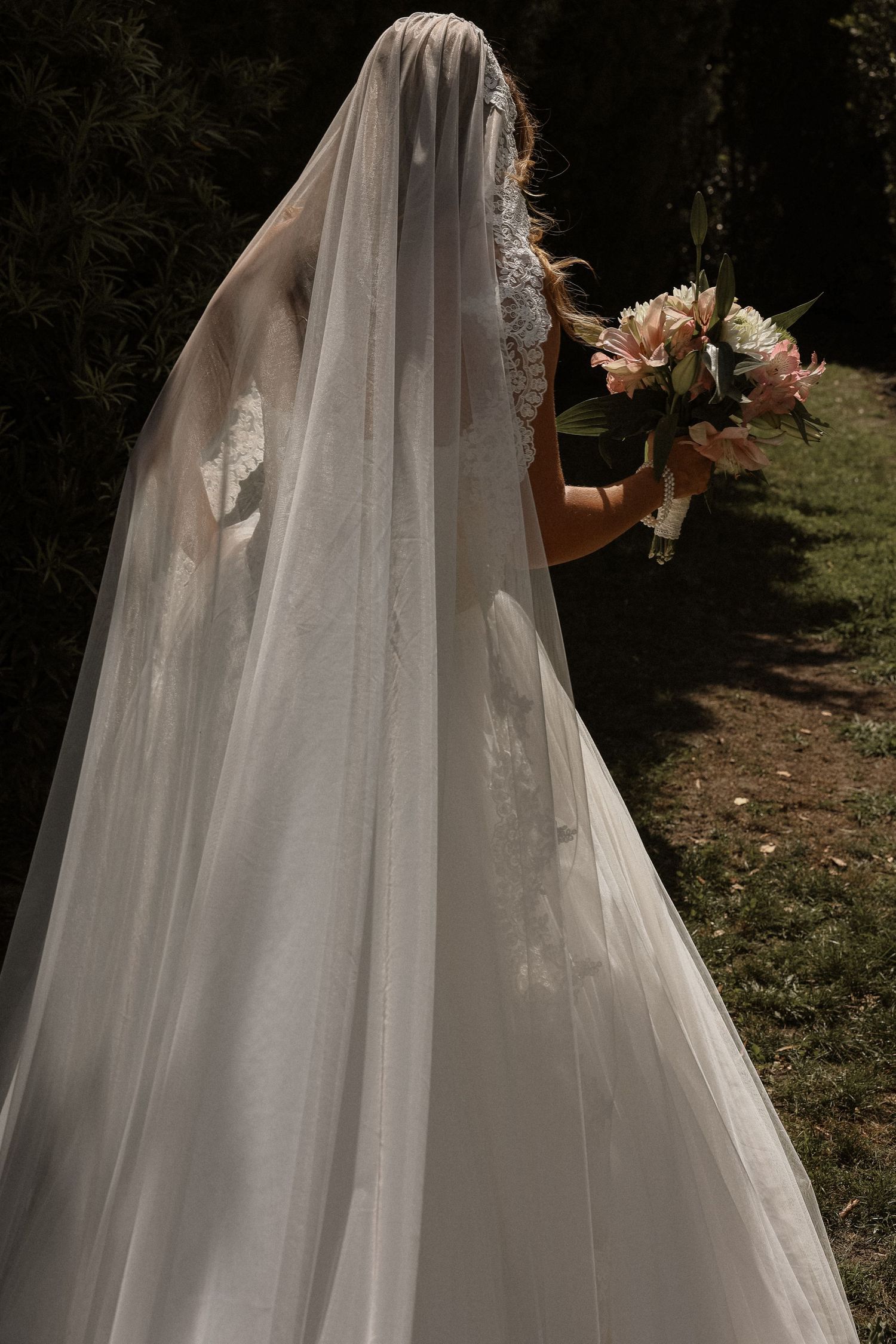 Bride in a flowing white wedding gown and long veil holds a pink bouquet, standing in a sunlit garden.
