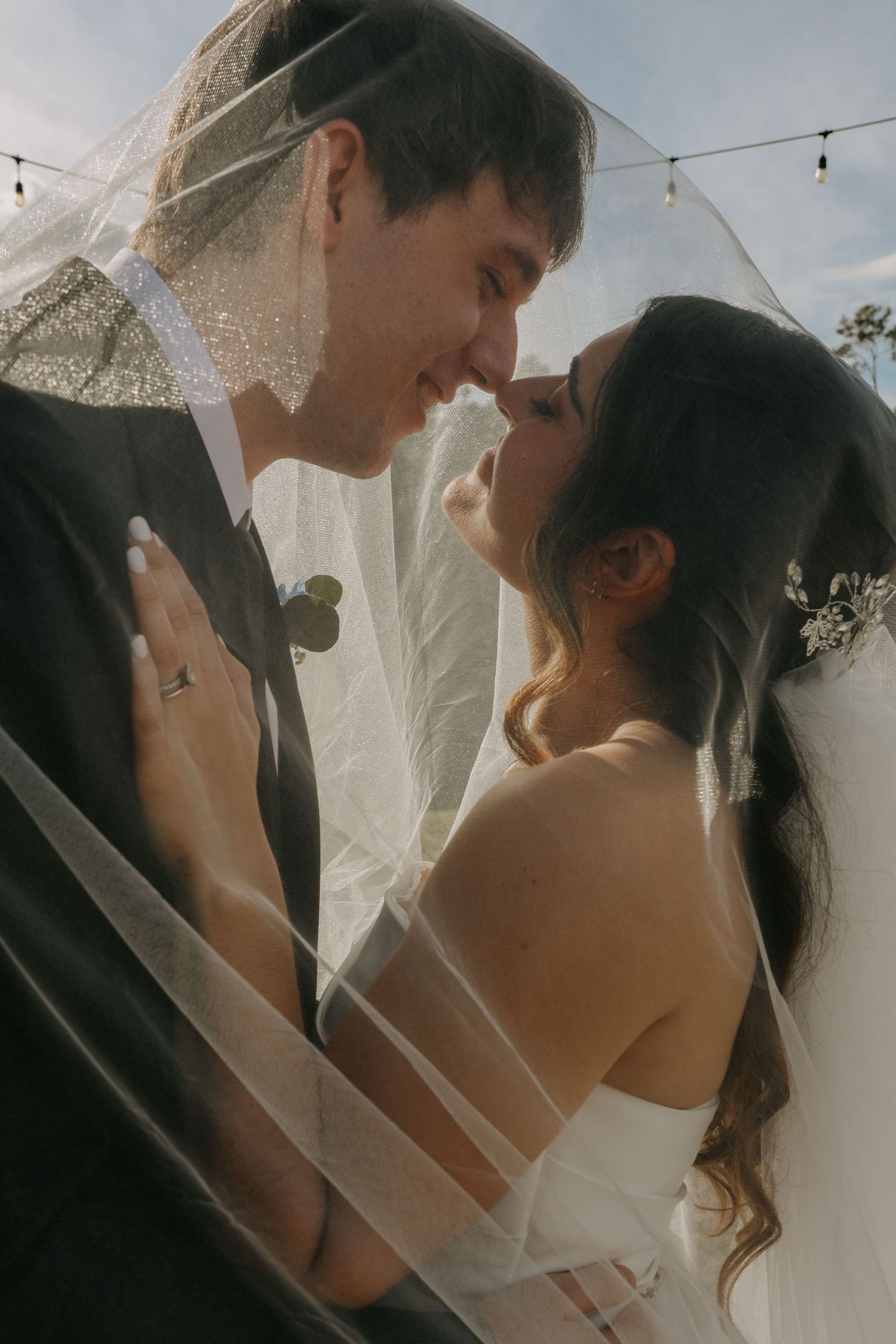 A bride and groom share a romantic moment under a flowing veil at their outdoor wedding ceremony.