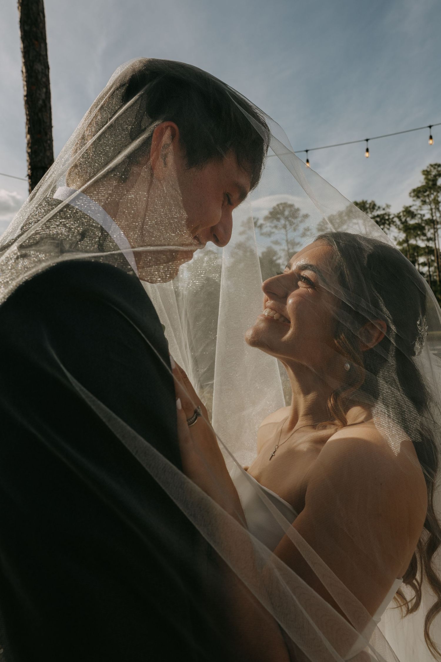 Bride and groom share romantic moment under wedding veil, smiling together outdoors with string lights in background.