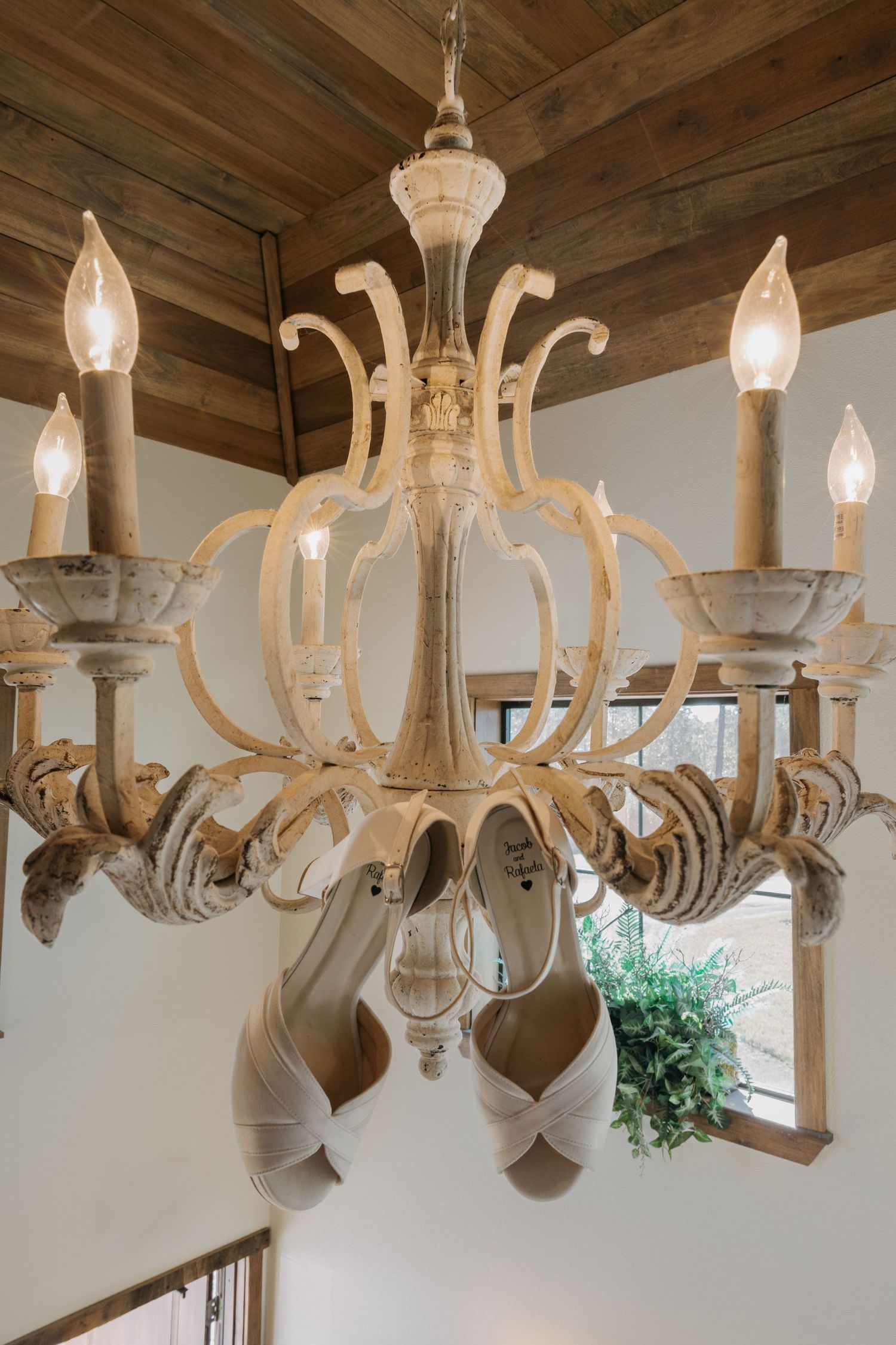 Vintage white chandelier with candle lights displaying bridal heels hung elegantly, set against rustic wood ceiling.