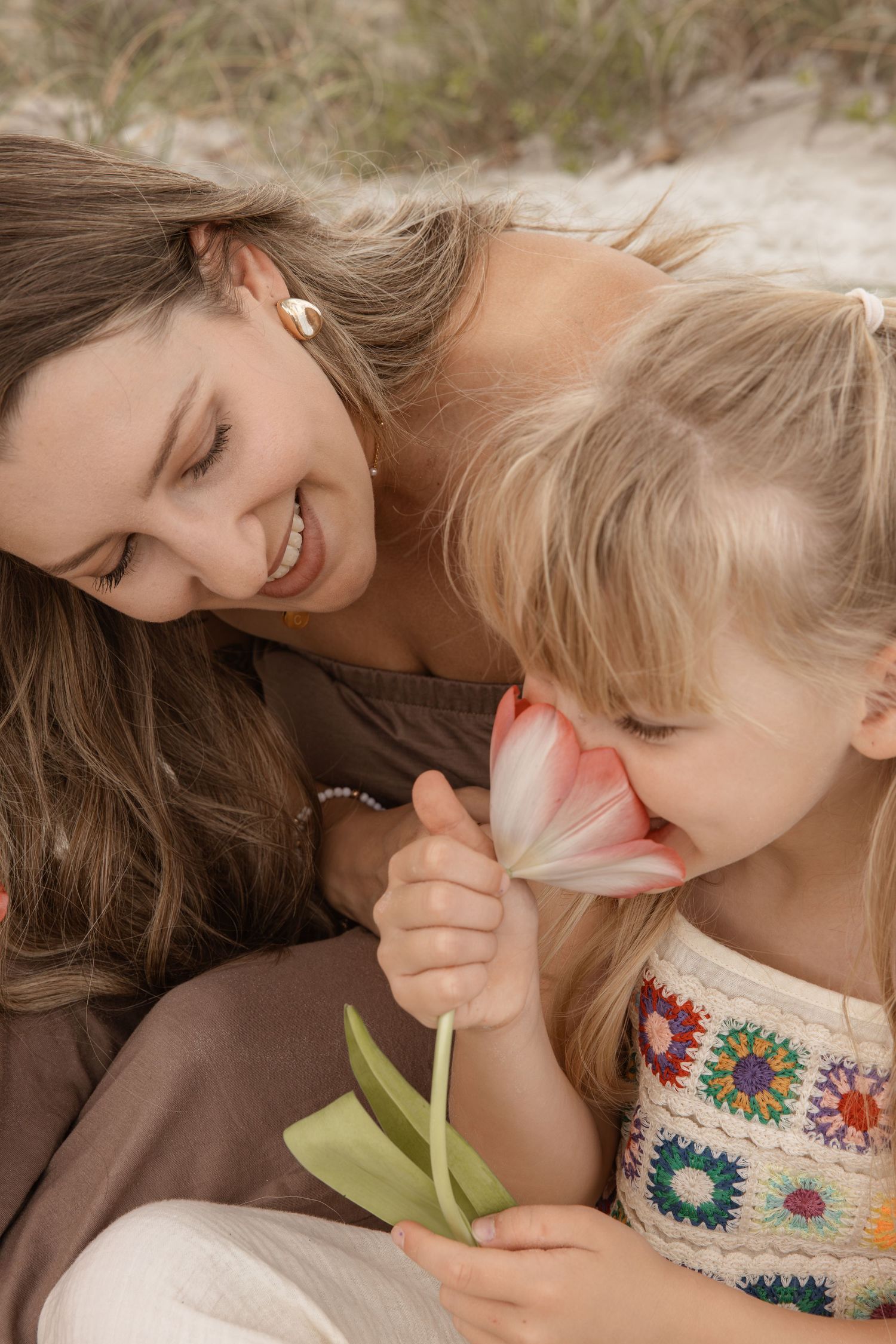 Smiling mother and blonde daughter smelling pink tulip flower together on sandy beach.