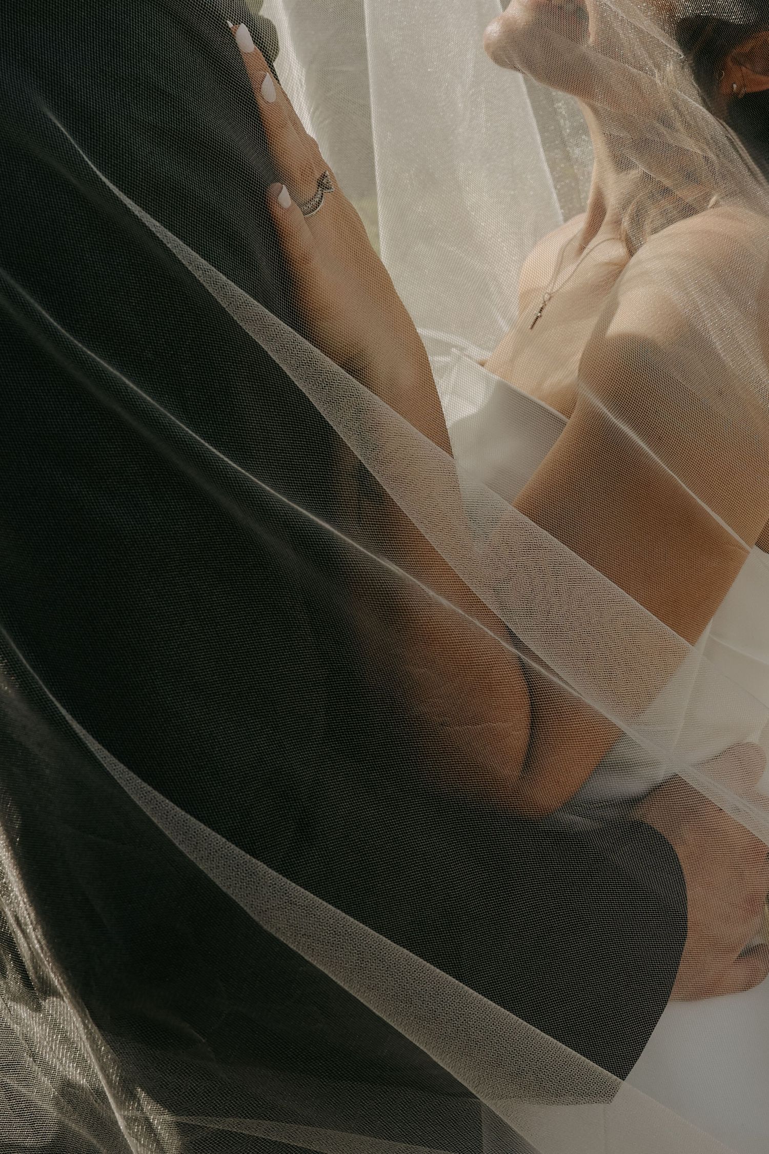 A bride and groom embrace under a flowing white wedding veil, captured in an intimate close-up detail shot.