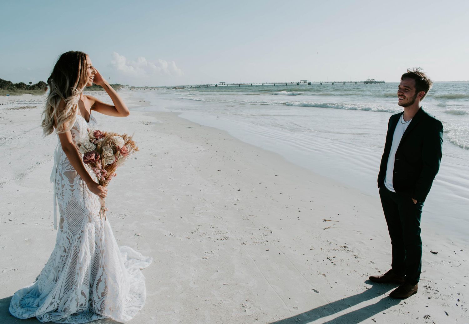 Bride in lace mermaid gown holds bouquet facing groom in black suit during romantic beach wedding ceremony.