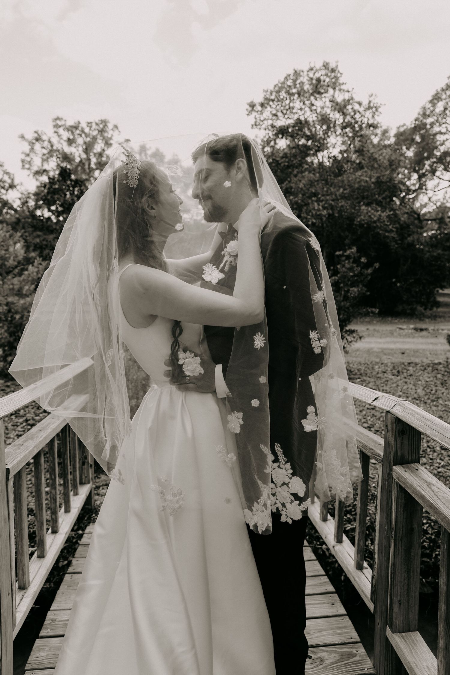 Black and white photo of bride and groom sharing romantic moment on wooden bridge, veil draped over both.