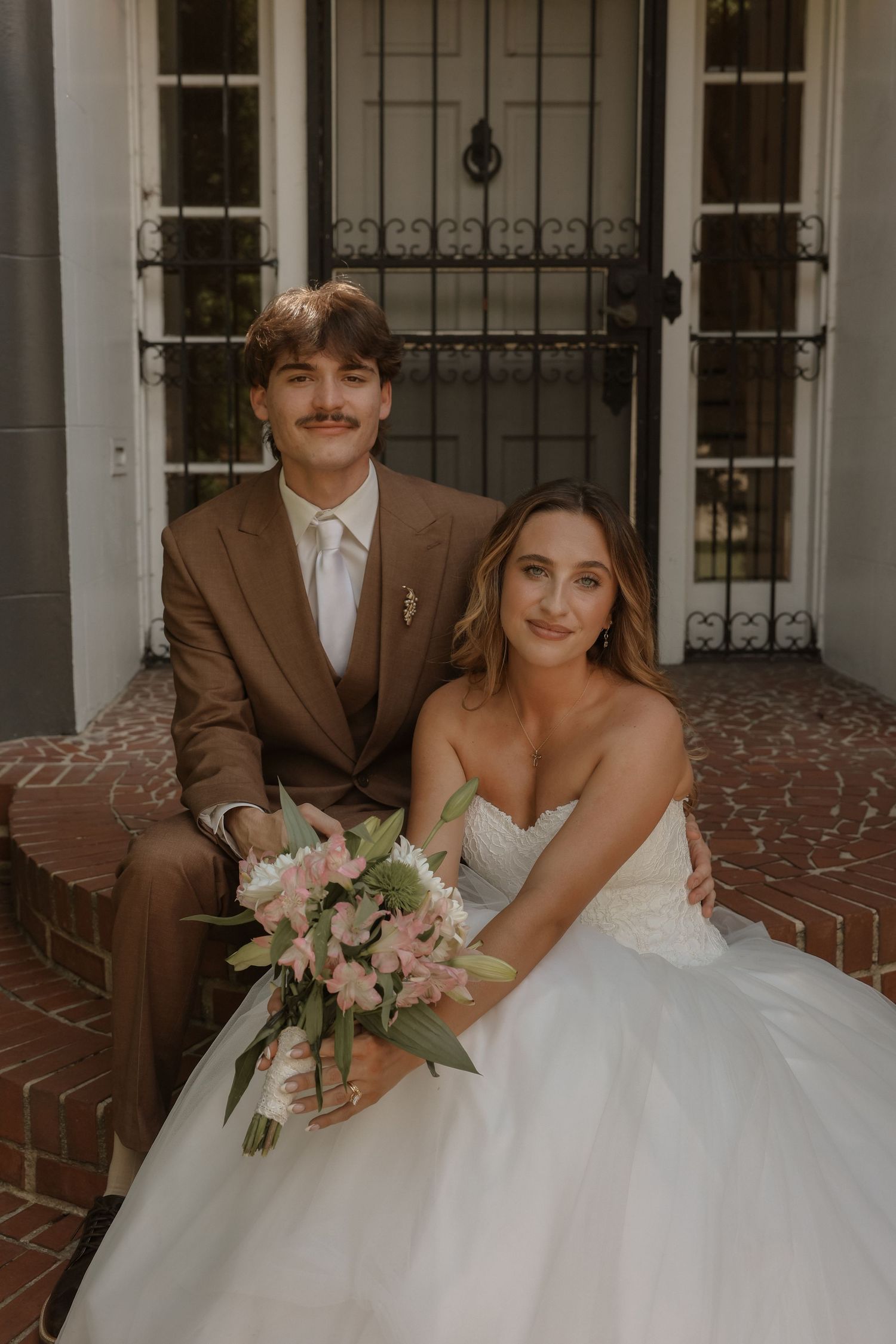 A couple in wedding attire sits on brick steps, groom in brown suit, bride holding pink floral bouquet in white gown.