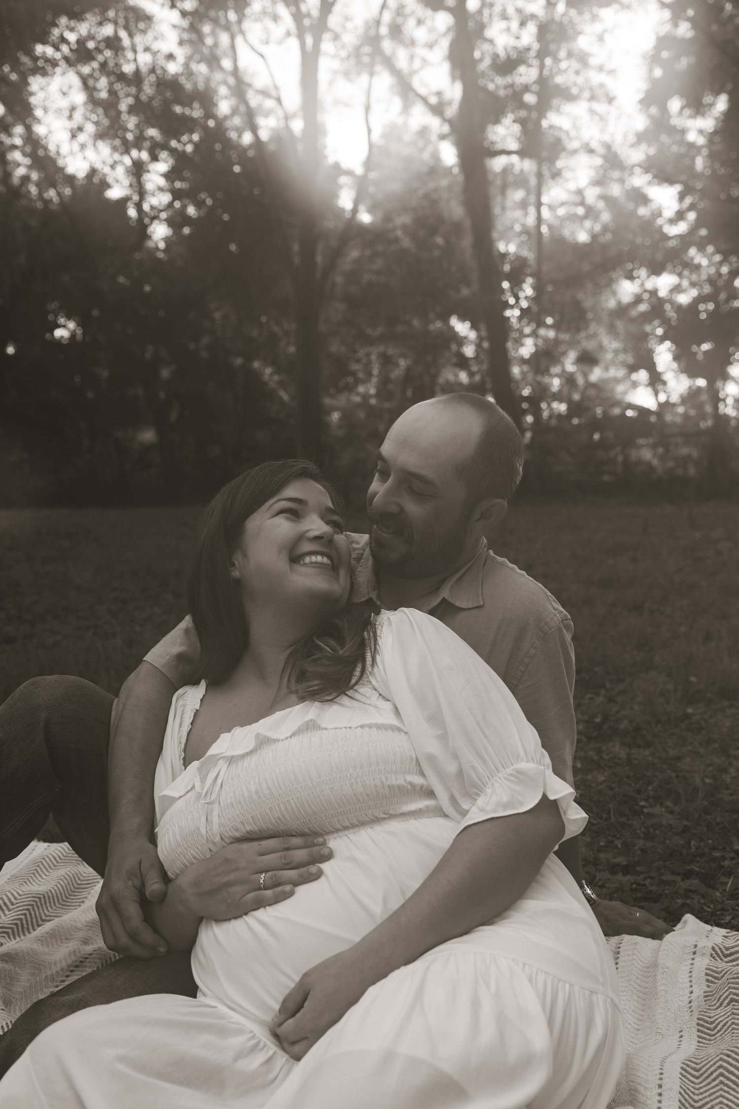 Black and white photo of a smiling pregnant couple sitting outdoors in a park, sharing a tender moment together.
