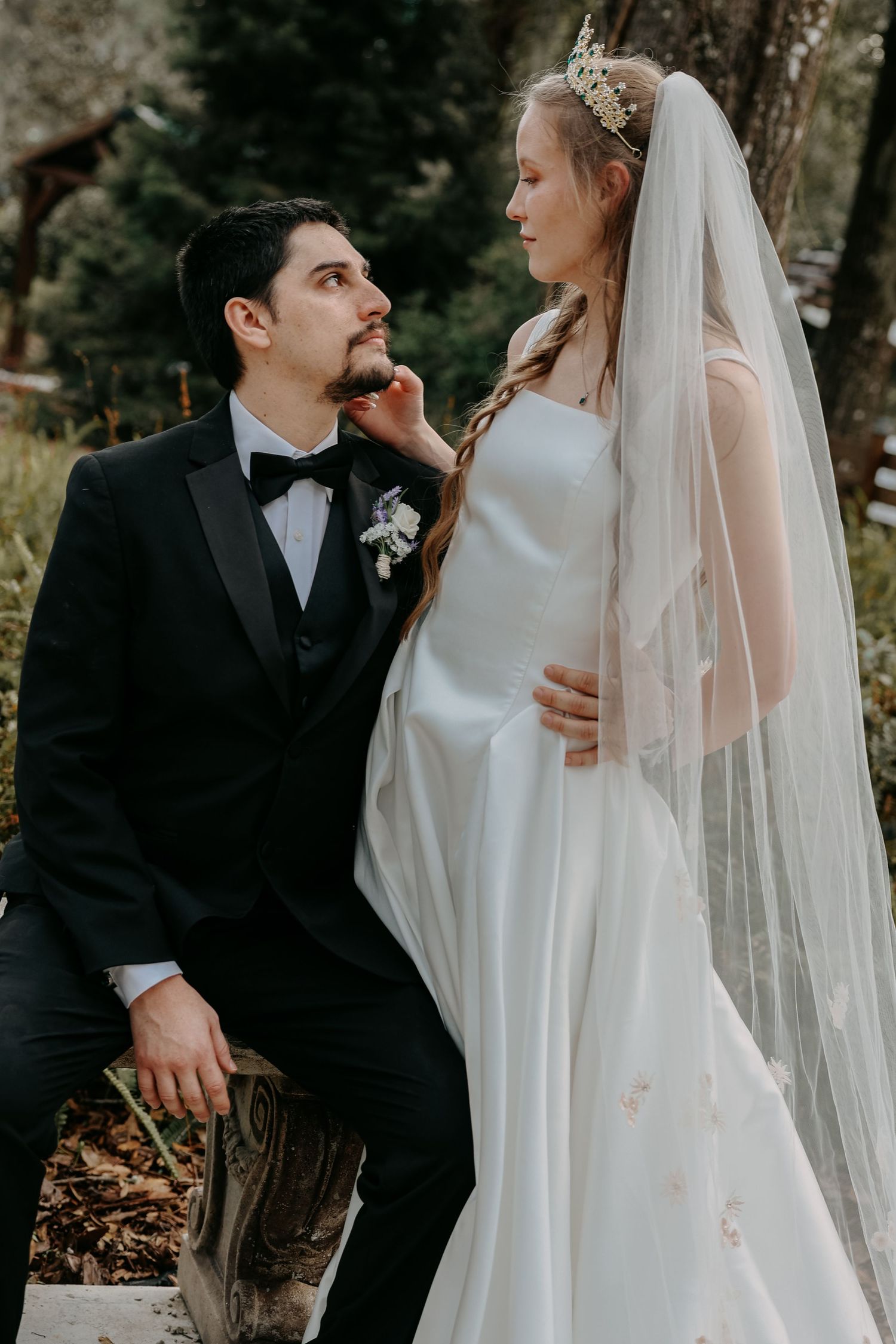 Bride in white gown and veil with groom in black tuxedo sharing a romantic moment outdoors.