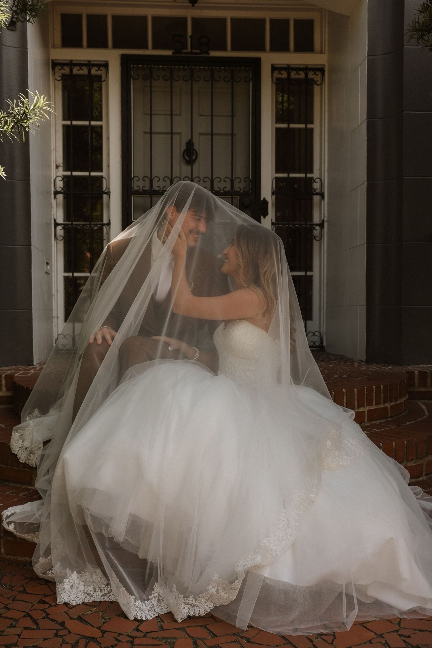 Bride and groom sharing intimate moment under sheer veil on brick steps of elegant building.
