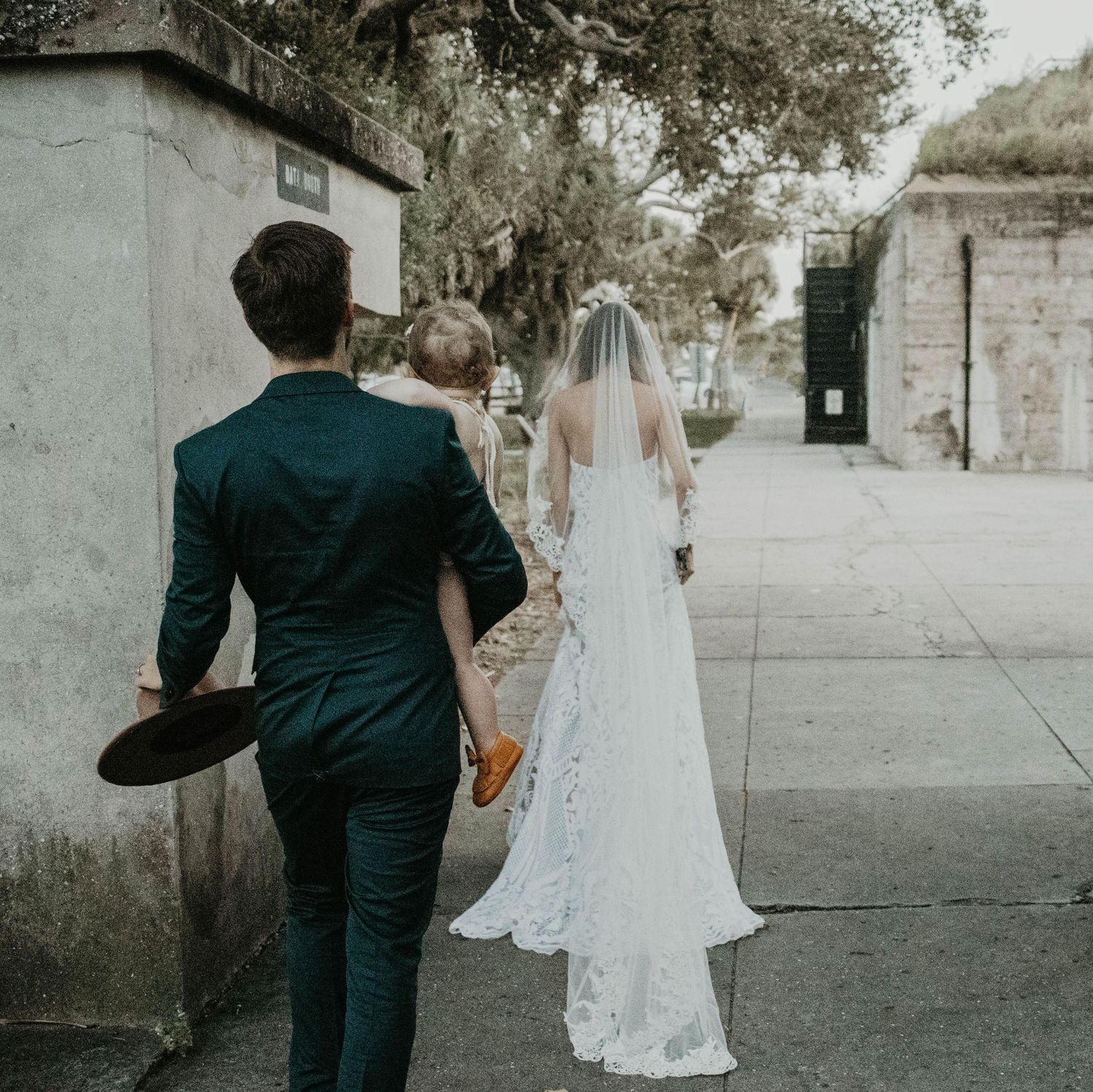 A bride in a lace wedding gown and veil walks alongside a groom in a dark teal suit on a scenic outdoor path.