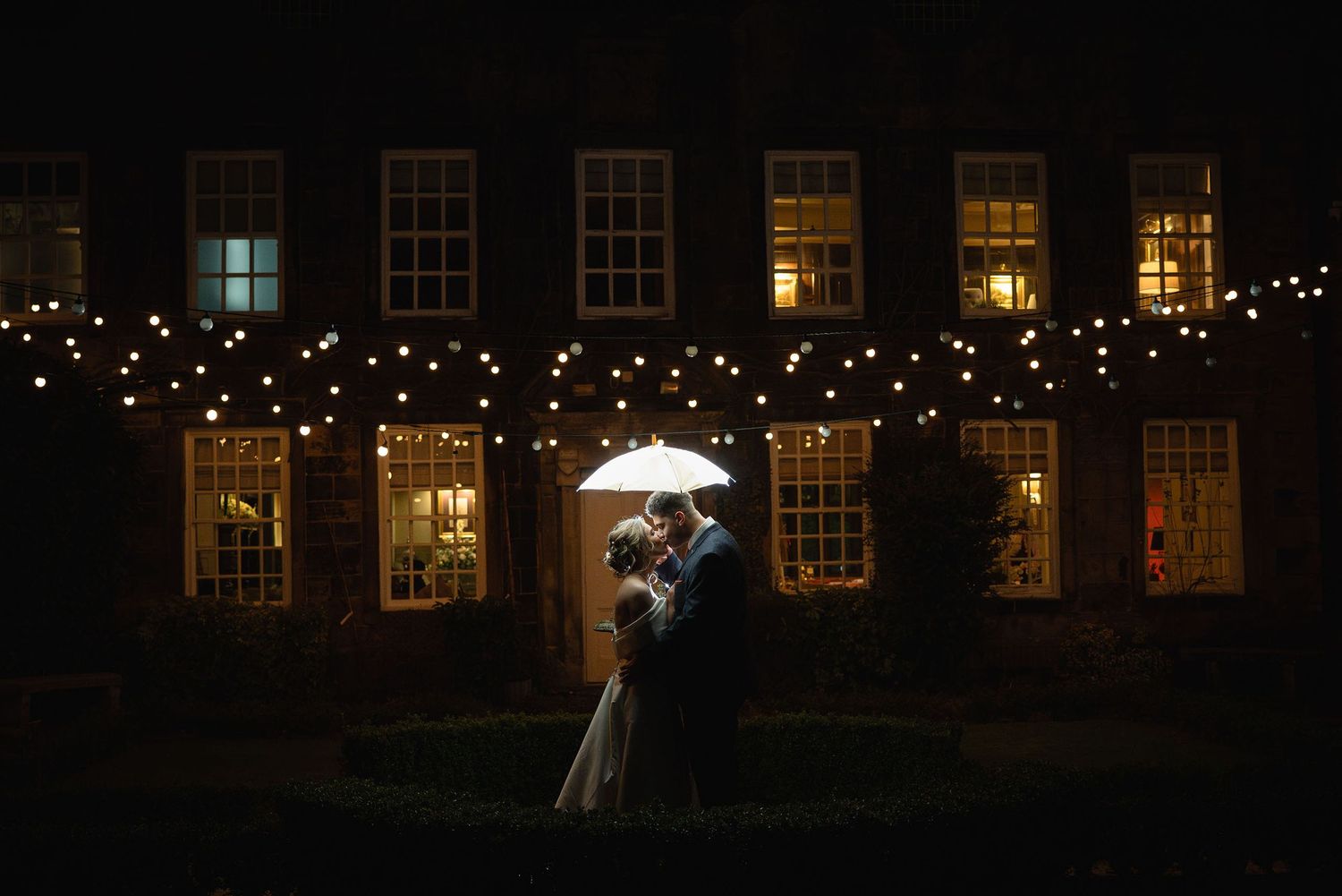 Off-camera flash portrait of the bride and groom kissing under an umbrella at night.