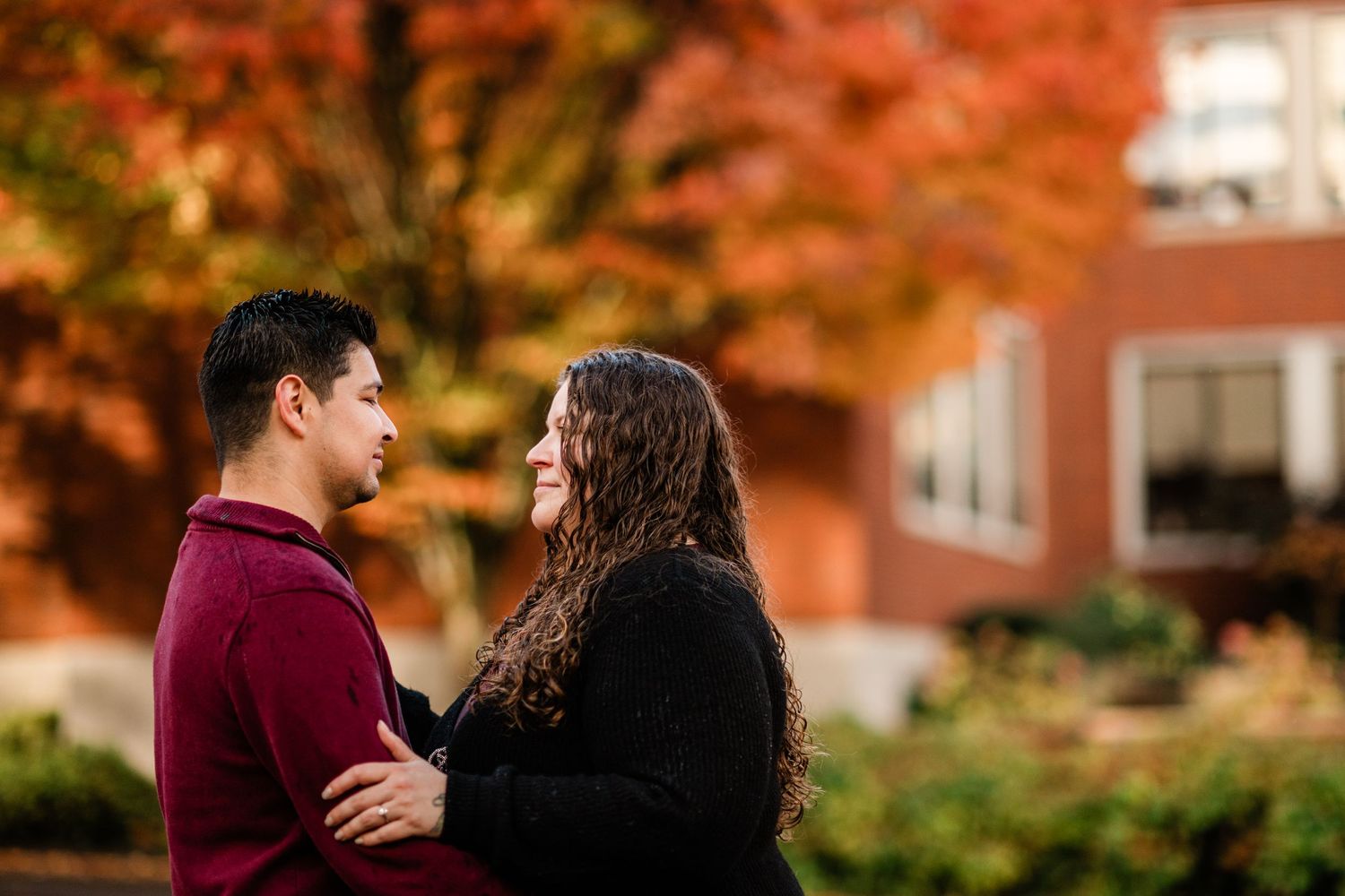 Couple gazing at each other with vibrant red autumn trees in the background during an engagement session.