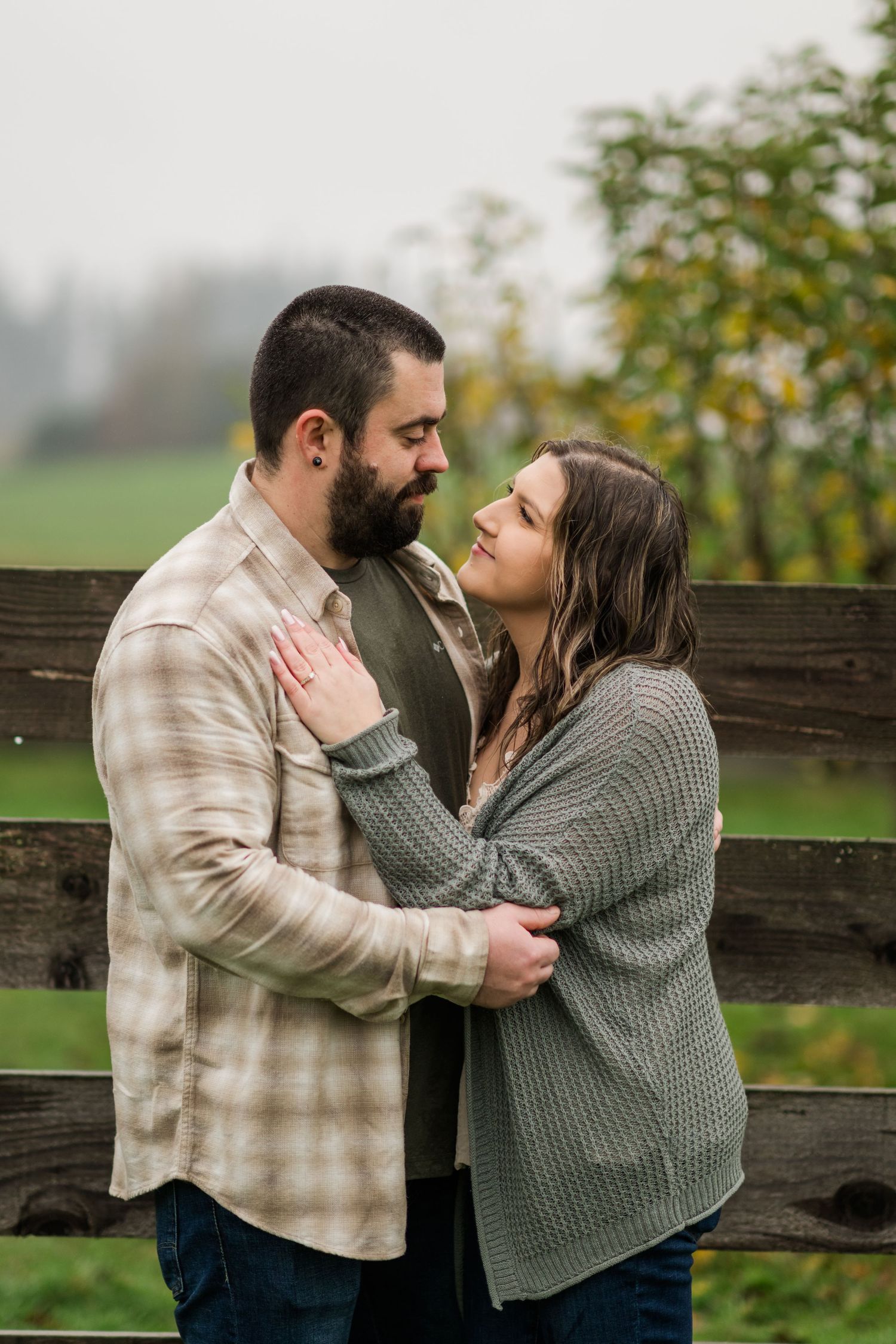 A bearded man and woman share a tender moment by a wooden fence on a farm.