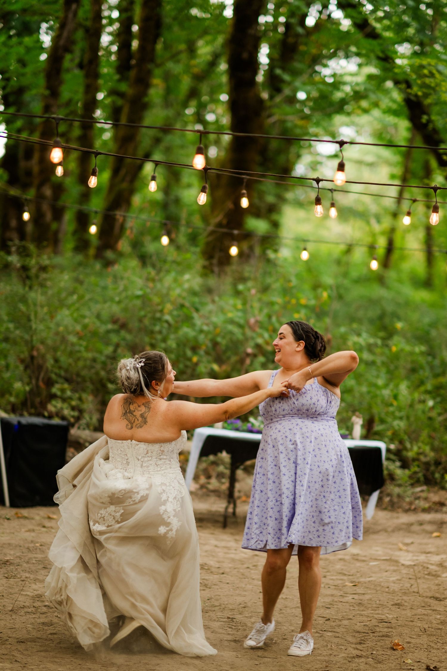 Bride in white gown dances joyfully with female guest under string lights at outdoor woodland wedding reception.