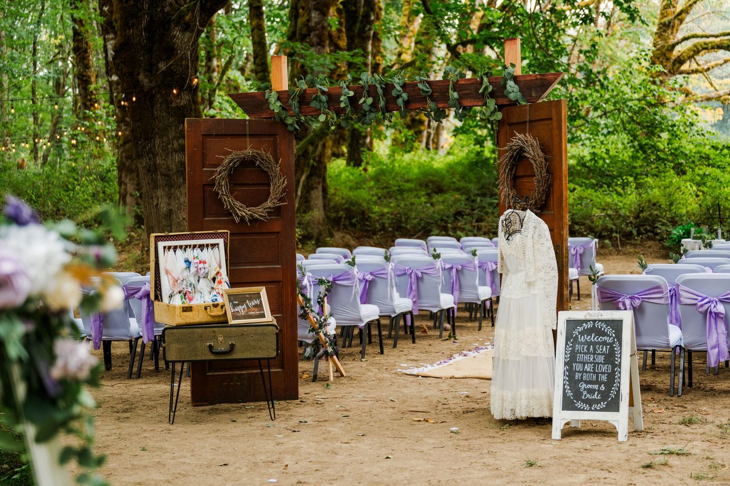 Woodland wedding venue with wooden arch, purple chairs, and white dress on display.