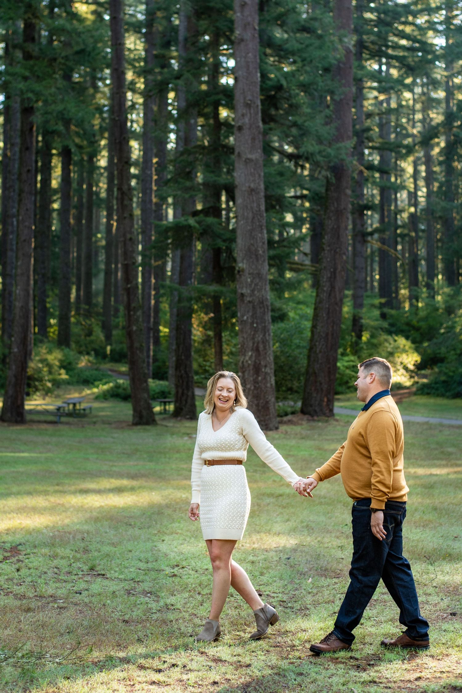 Playful couple in white dress and brown sweater dancing in sunny forest clearing.