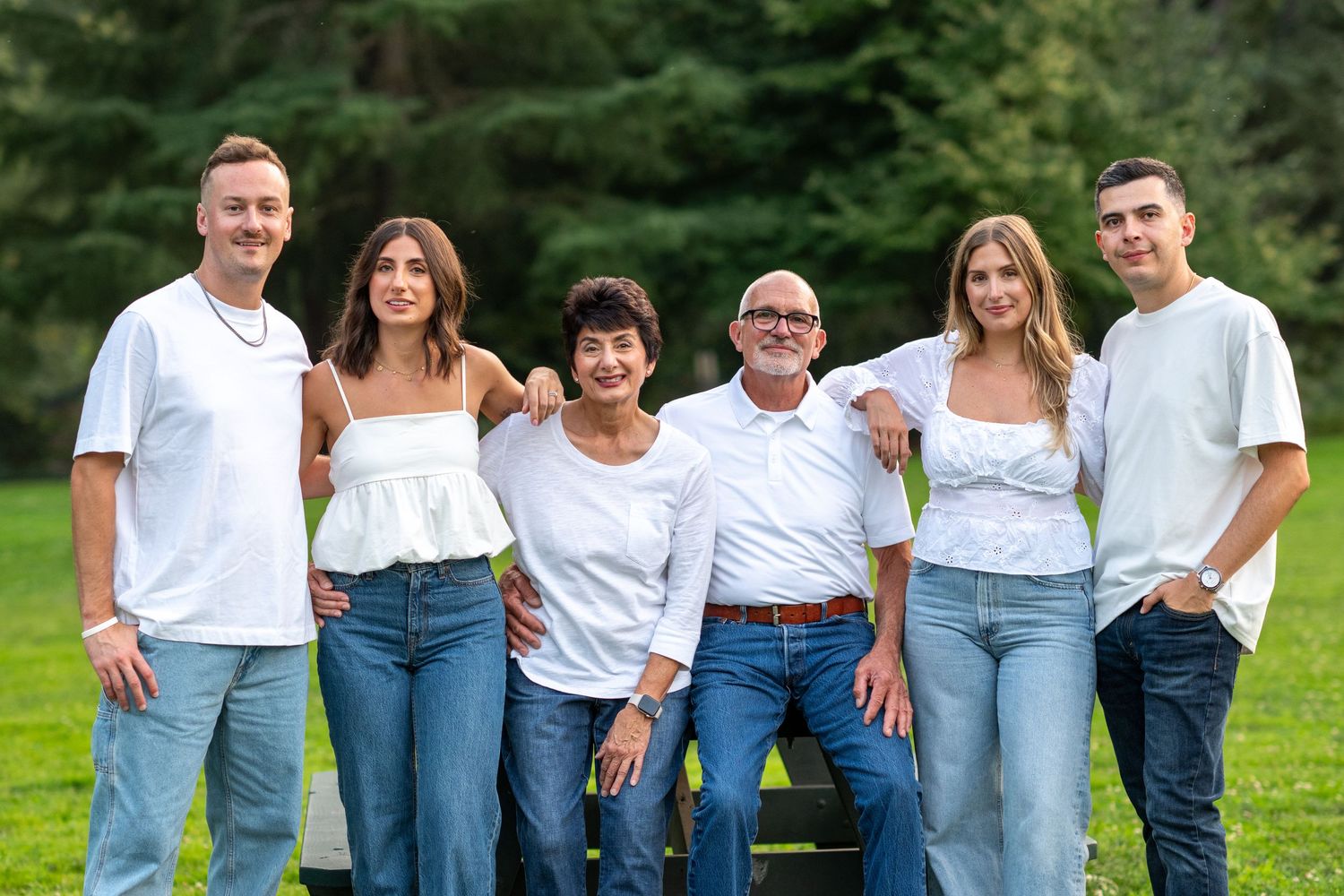 Family of six in white tops and jeans posing together on a bench in a park.