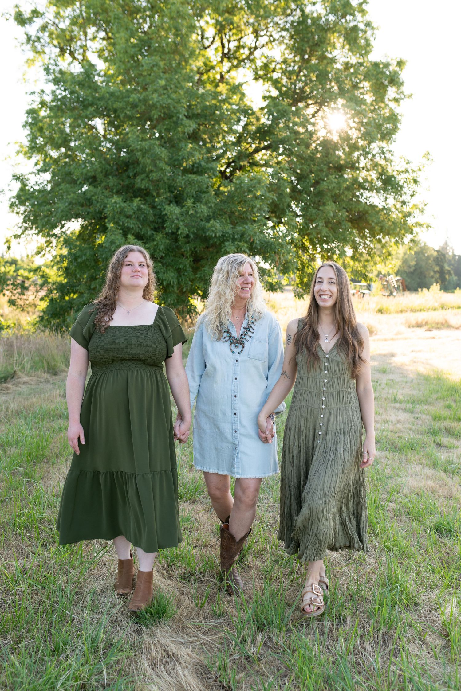 Three women in green and blue dresses walking hand in hand through a sunlit field near a large tree.
