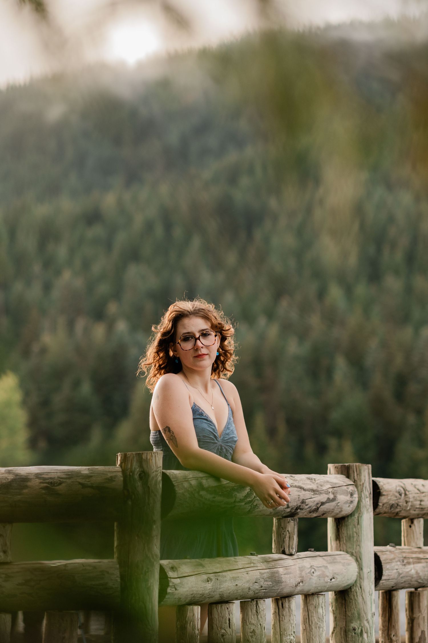 Woman with curly red hair and glasses leans on a rustic wooden fence with a forested mountain backdrop.